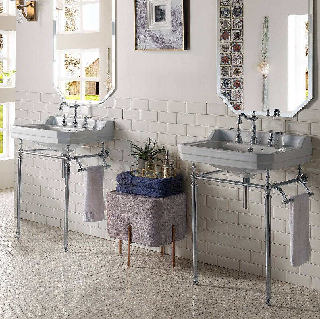 Two stone-colored ceramic console sinks with chrome metal legs and faucets mounted on a white subway-tile wall, flanked by mirrors; an upholstered ottoman with folded navy towels and a decorative tray sits between them.