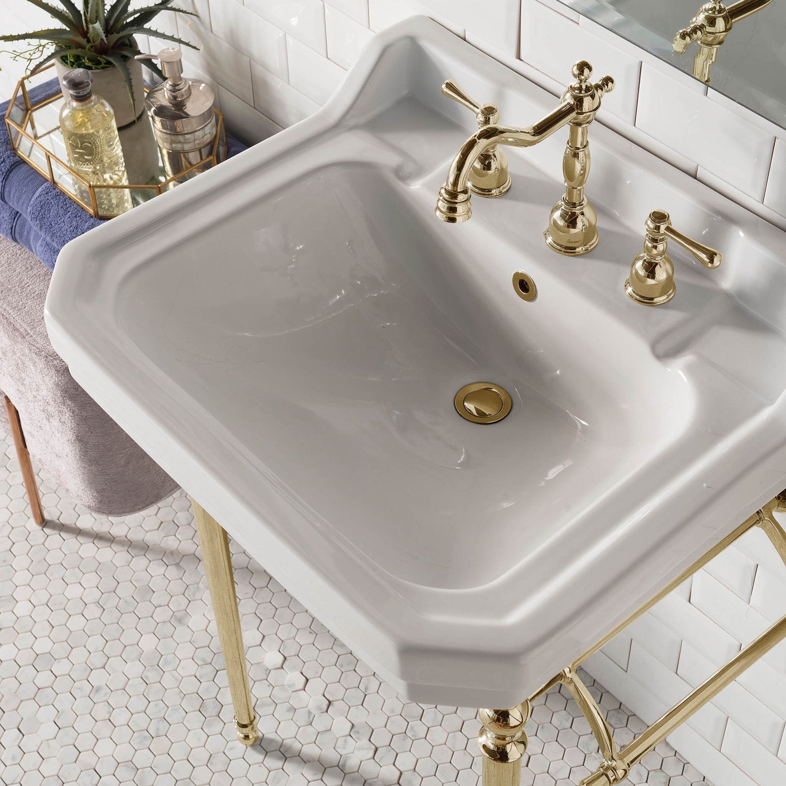 White ceramic single-console sink with polished brass widespread faucet and matching brass metal legs, shown against white subway tile and hexagon floor tile with folded towels and a decorative tray.