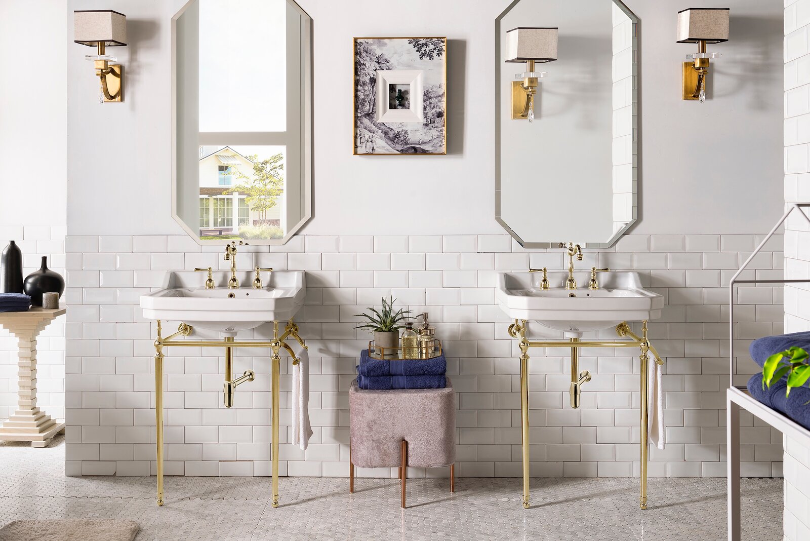 Two white ceramic console sinks with polished brass legs and faucets mounted on a white subway-tile wall, each beneath a faceted mirror and brass sconce; a stone-colored stool with folded blue towels and tray sits between them.