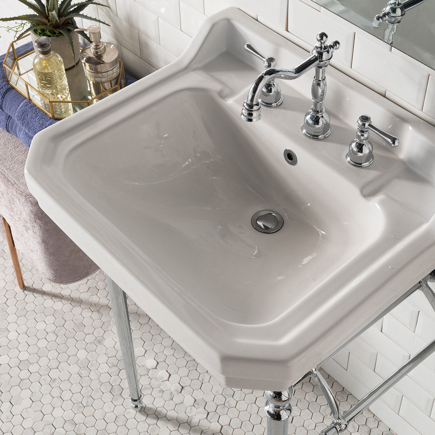 Angled top-down view of a stone-colored ceramic console sink with chrome widespread faucet, pop-up drain and metal legs, staged with folded towels and a decorative tray on white hex-tile floor.