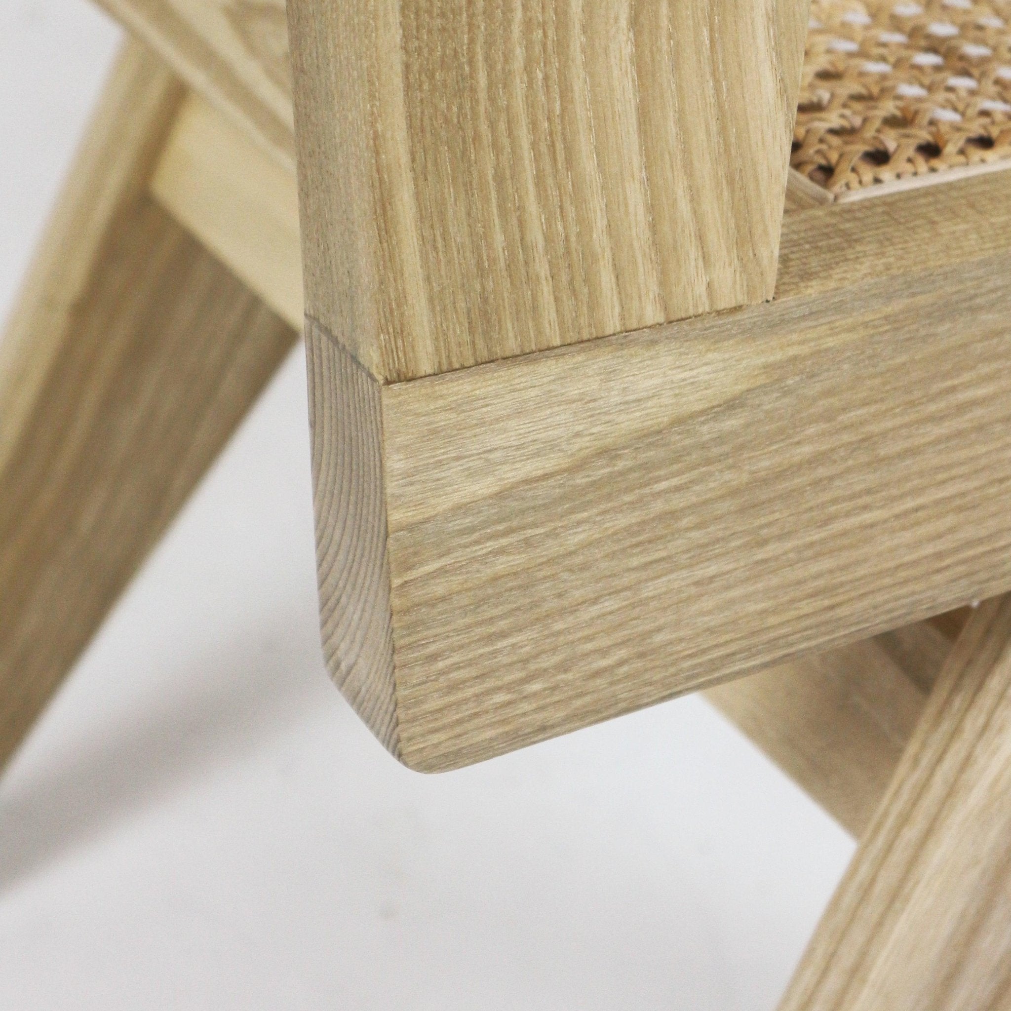 Close-up of an armless side chair's solid natural wood joint and frame in a light blonde finish, showing a rounded corner detail and a portion of the woven cane seat.