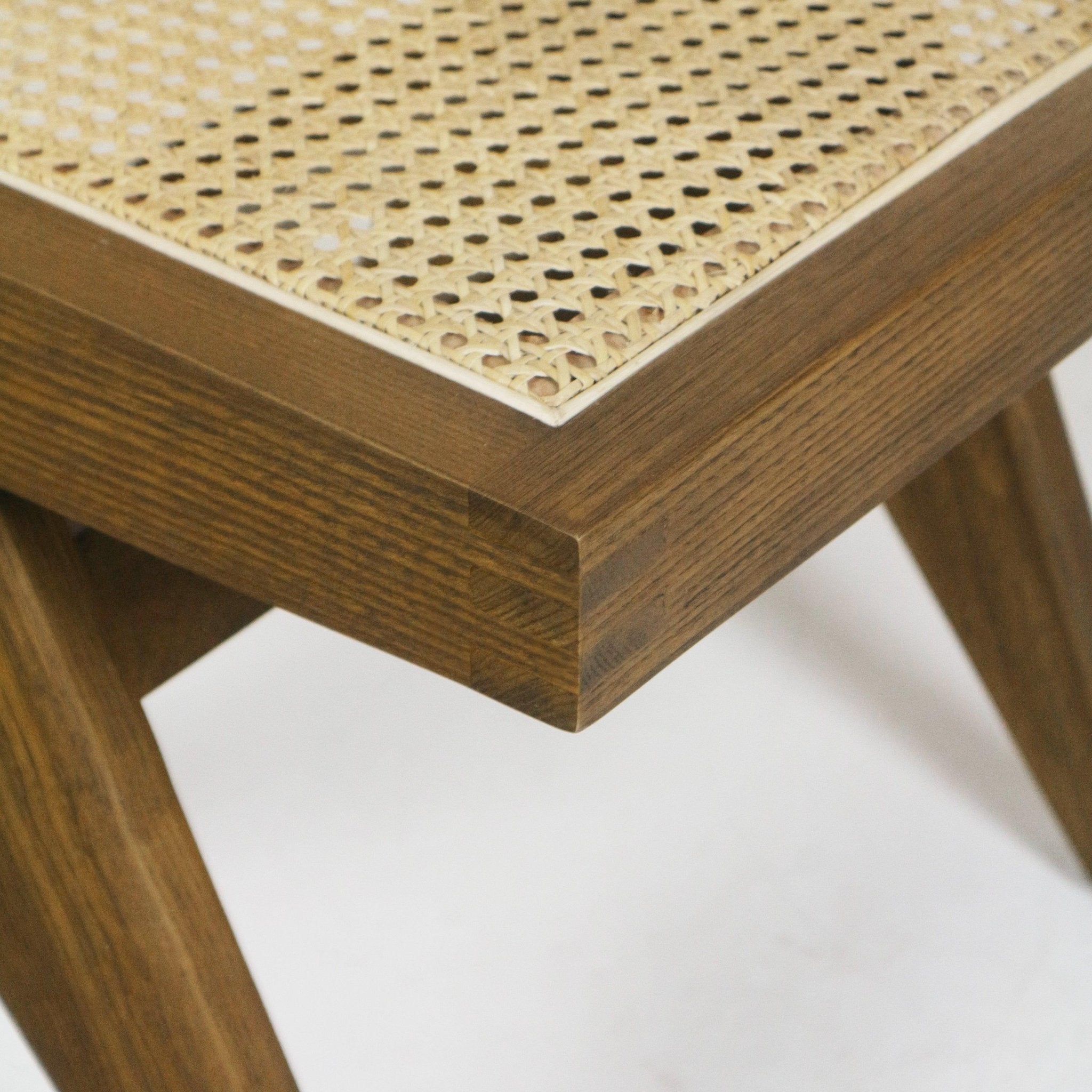 Close-up of a warm teak solid-wood chair corner showing woven natural cane seat, visible finger-joint detail, light edge trim and a tapered leg on a modern armless dining side chair.