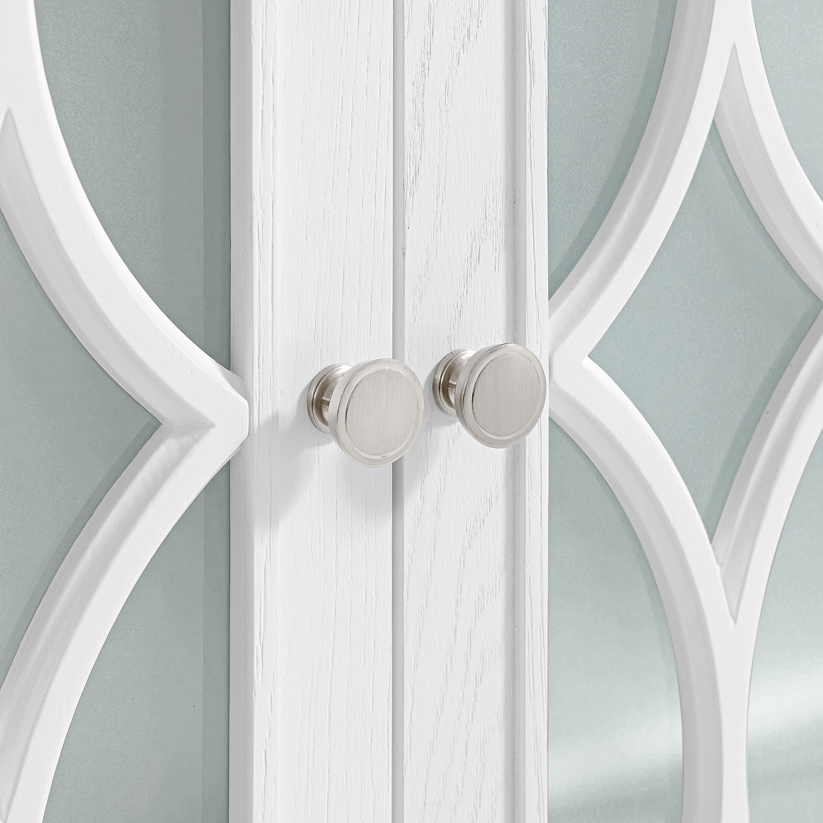 Close-up of modern white solid wood vanity doors with decorative arched trim and two round brushed-nickel knobs against a pale stone-toned mirrored background.