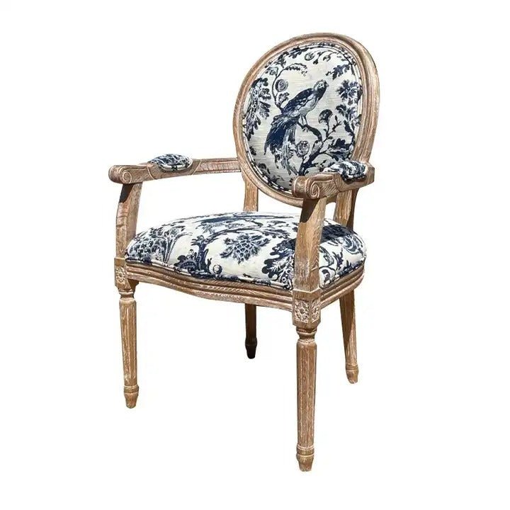 French-style dining armchair with a carved teak frame and oval back, upholstered in cream linen with blue toile bird-and-floral print, padded arm rests and a cushioned seat on turned fluted legs.