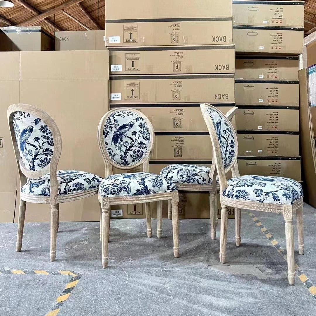 Four French-style dining chairs with weathered teak frames and blue-and-white toile linen upholstery, oval backs and carved legs, arranged on a concrete floor in front of stacked cardboard shipping boxes.