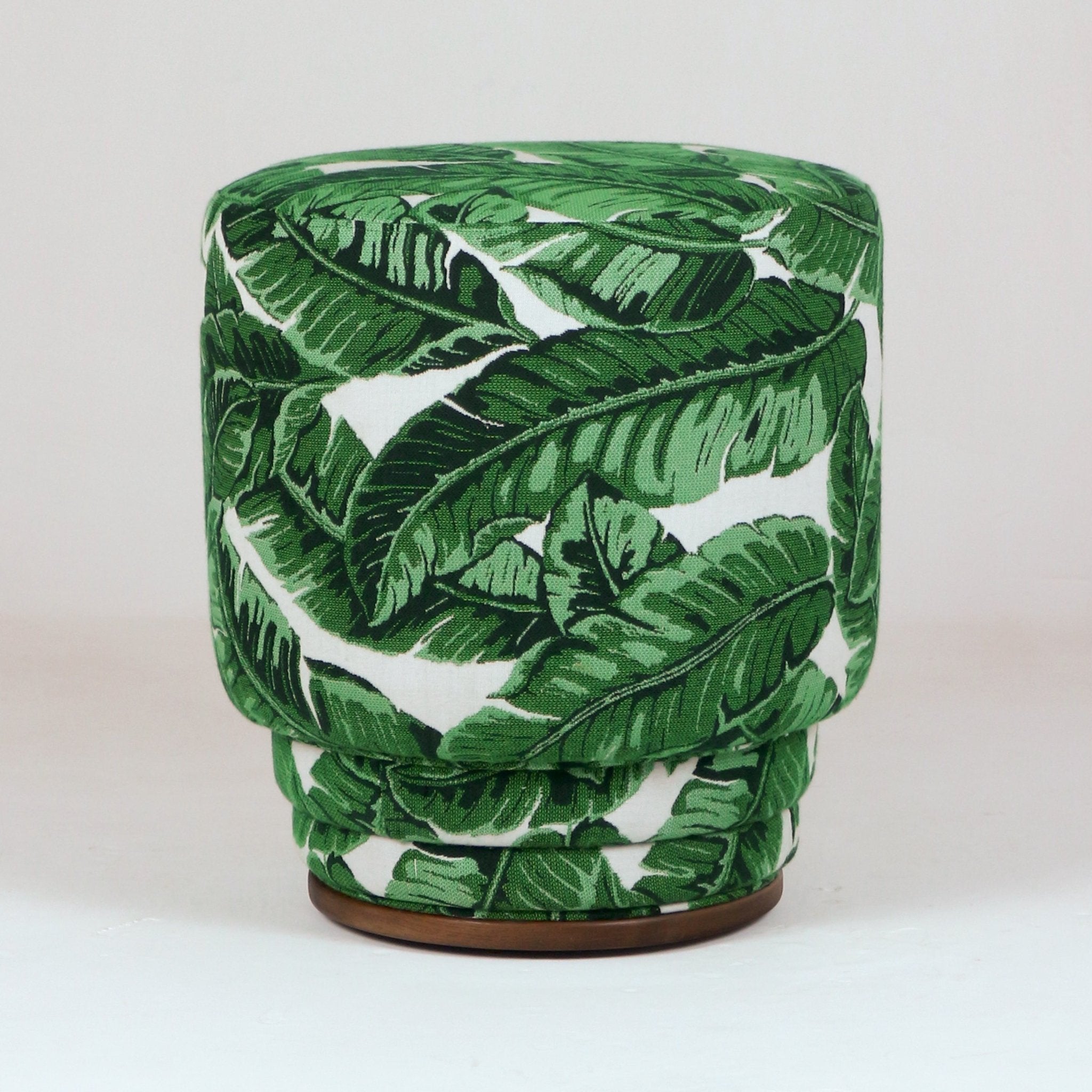 Round upholstered ottoman in green tropical leaf print fabric on a white background, featuring a stacked cushioned silhouette and low wooden base for a modern indoor or outdoor seating accent.