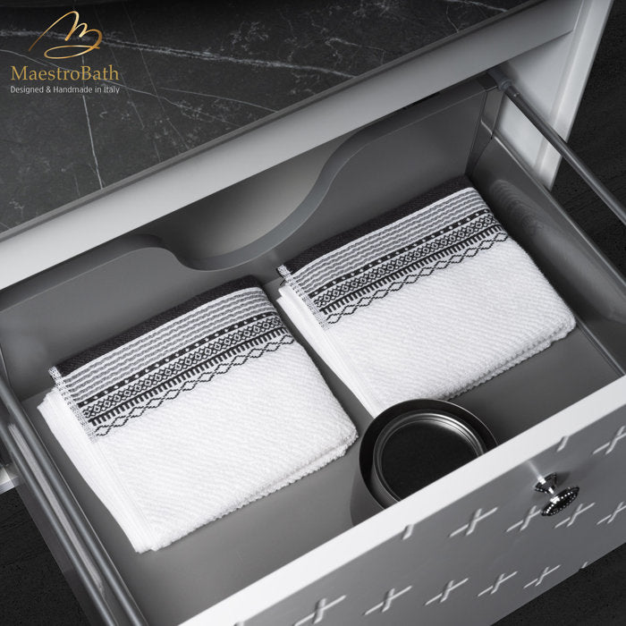 Open stone-top bathroom vanity drawer with gray interior holding two folded white towels with black embroidered borders and a small black round container; white drawer front shows decorative cross pattern and chrome knob.