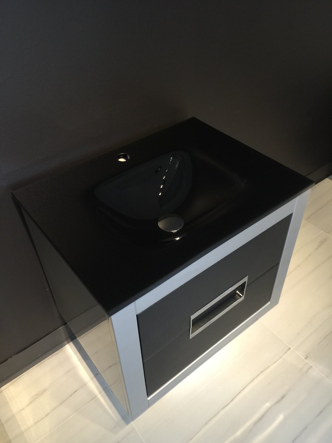 Freestanding modern single-sink bathroom vanity with a black stone top and integrated basin, solid wood frame, single gray drawer with chrome rectangular pull, photographed against a dark wall on light tile flooring.