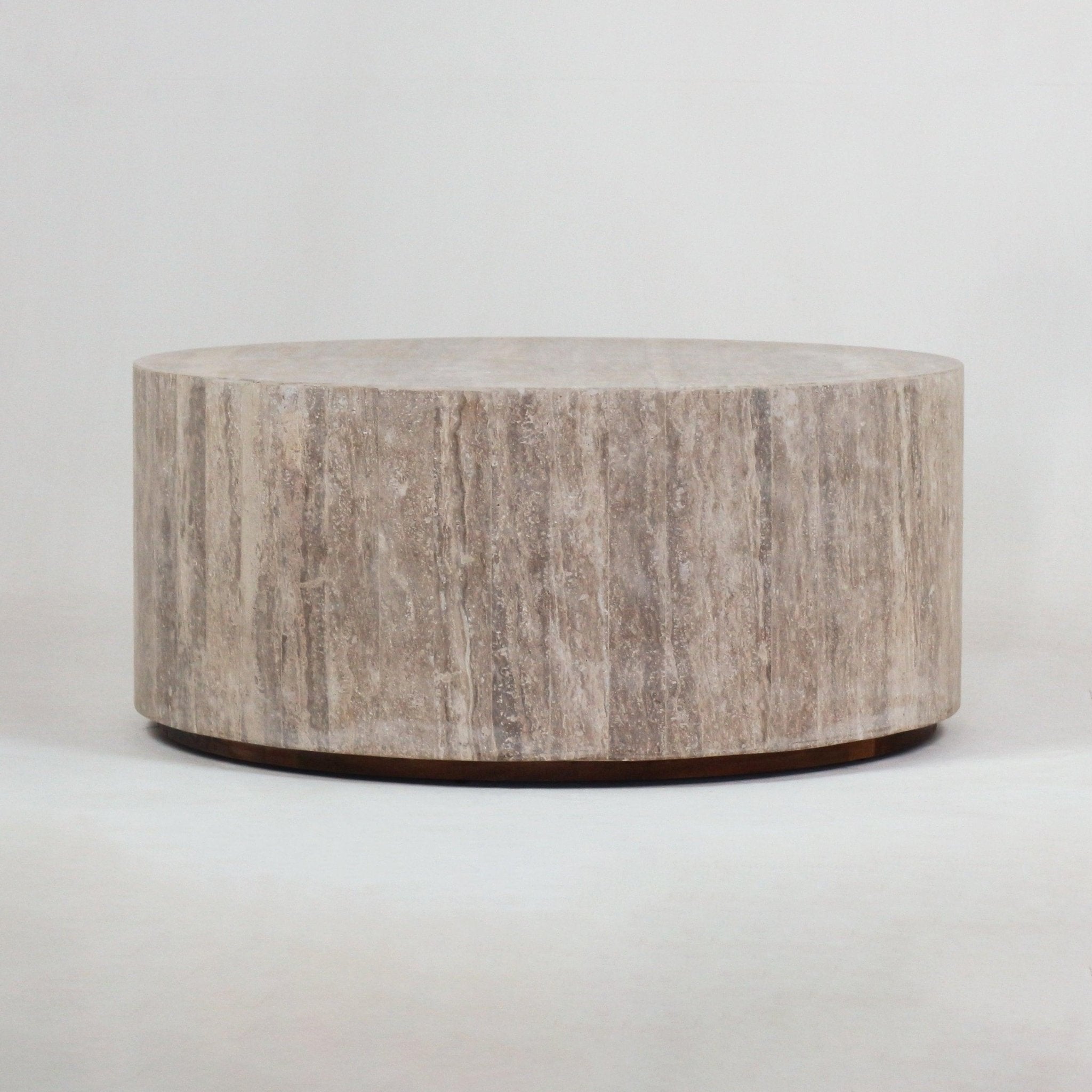 Low-profile round travertine coffee table with cylindrical drum shape, natural beige stone veining and a slim dark recessed base on a neutral studio background.