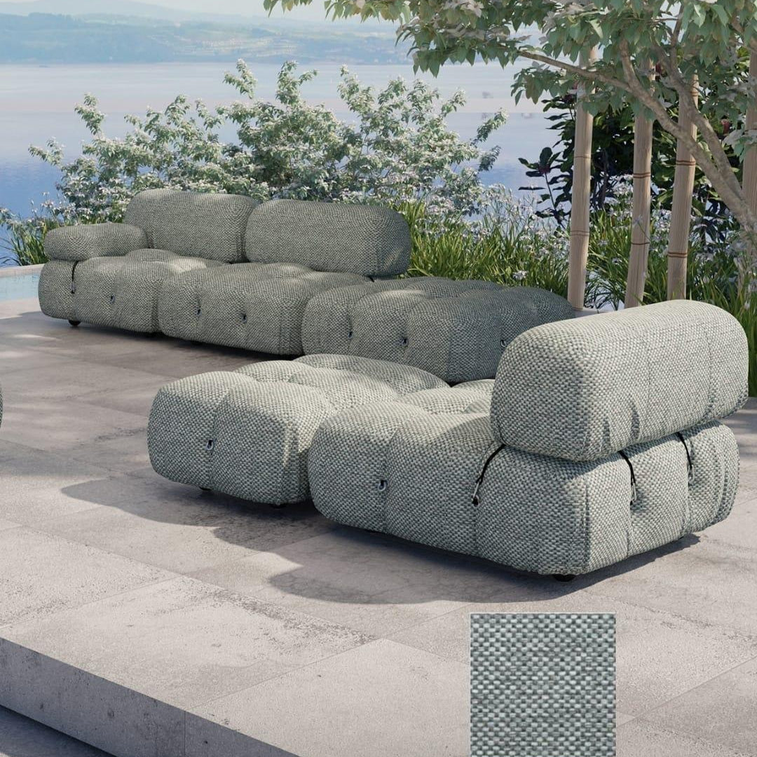 Coastal-modern outdoor modular sofa and matching ottoman upholstered in textured woven fabric in muted sage-gray, with tufted rounded cushions, shown on a stone patio beside greenery and a sea view.