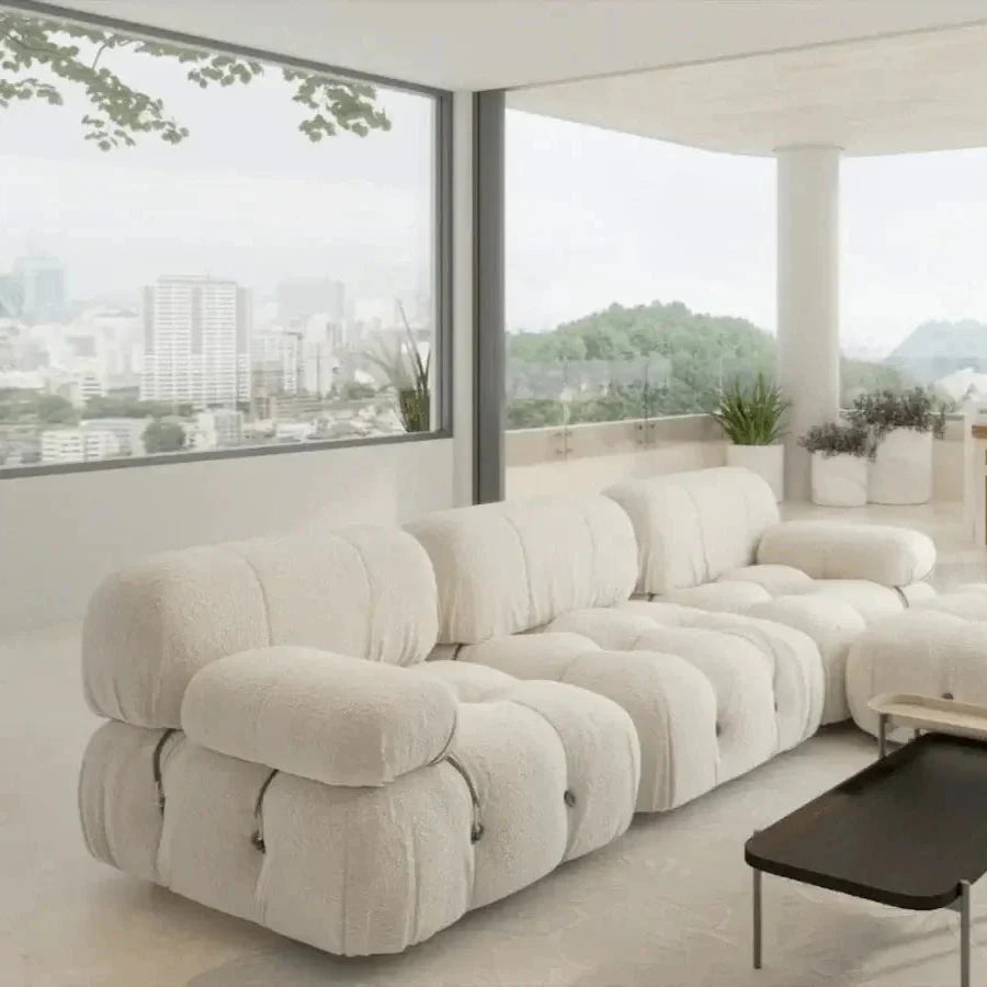 Cream boucle fabric three-seat modular sofa with rounded tufted cushions and low arms, shown in a modern living room by floor-to-ceiling windows with potted plants and a black coffee table on a neutral rug.