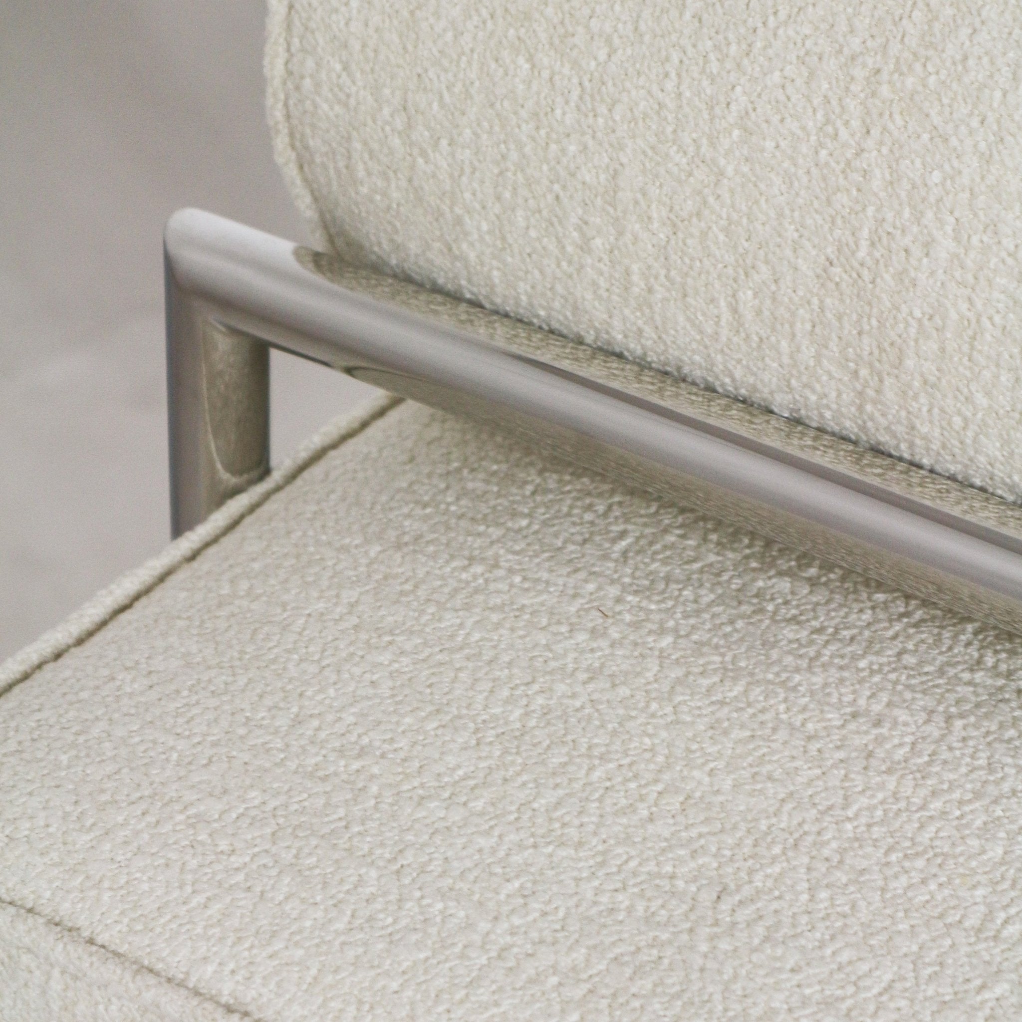 Close-up of cream boucle upholstery on an end-of-bed bench showing textured fabric seat and back with a brushed stainless steel frame rail and metal arm detail.