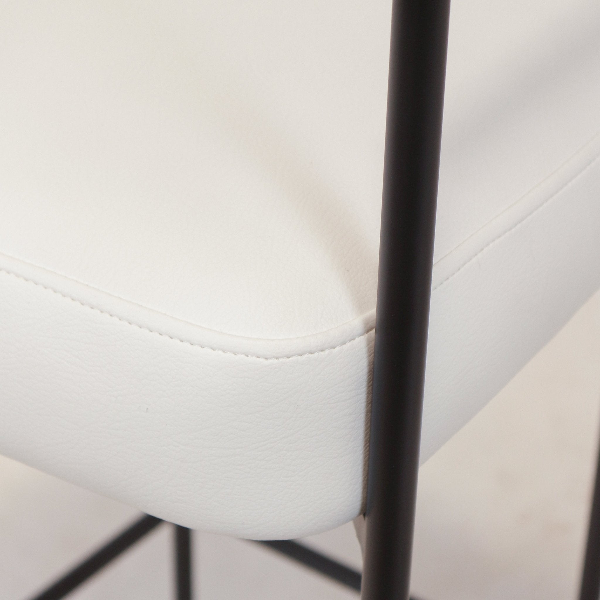 Close-up of a white faux leather seat cushion with neat topstitching and rounded edge, shown alongside a slender matte black metal leg and crossbar from a modern counter/bar stool.
