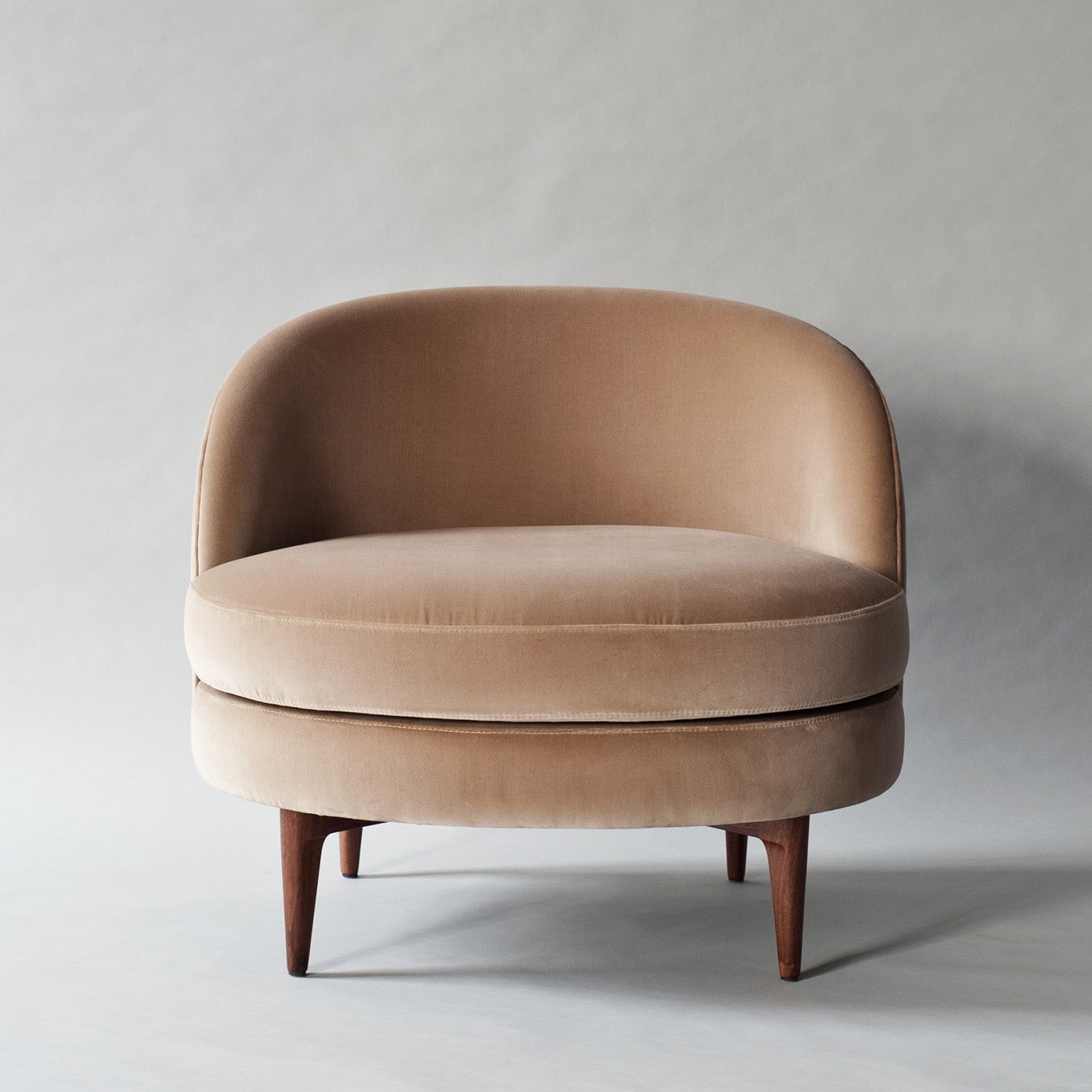 Contemporary blush velvet accent chair with a rounded barrel back, thick cushioned seat and tapered solid wood legs, photographed on a neutral studio background.