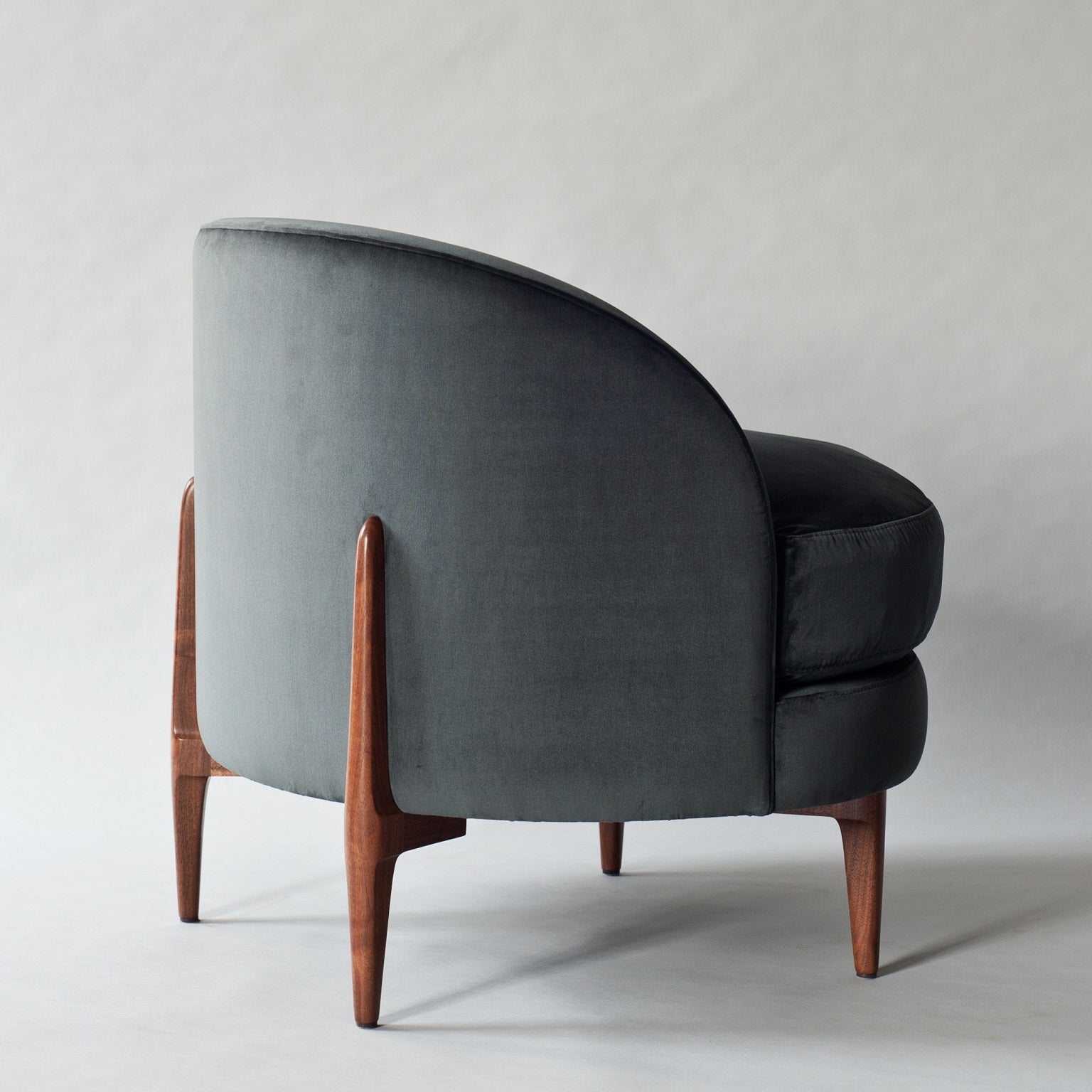 Rear three-quarter view of a modern charcoal gray velvet accent chair with a curved barrel back, plush rounded seat cushion and tapered solid-wood legs in a warm walnut finish.
