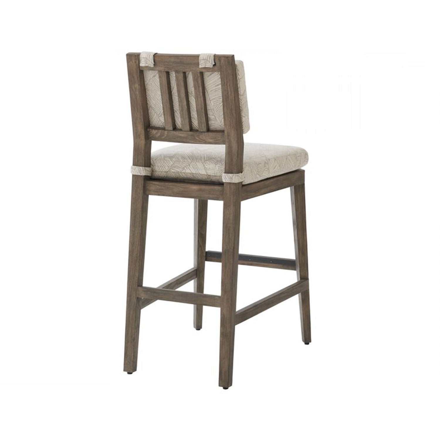Rear three-quarter view of an armless teak outdoor bar stool with a beige patterned upholstered seat and low slatted back on a natural teak wood frame with square legs and a built-in footrest.