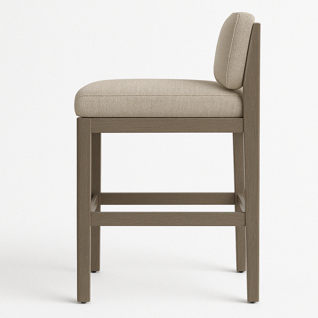 Side view of an armless teak bar stool with a beige upholstered cushion and low padded back, natural teak square legs and integrated footrest in a coastal contemporary outdoor style.