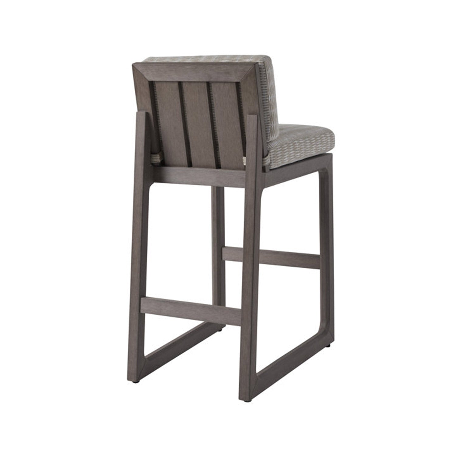 Rear three-quarter view of an armless teak outdoor bar stool with a grey teak frame and beige woven fabric seat and back cushion, showing vertical slatted back, open-leg frame and built-in footrest.