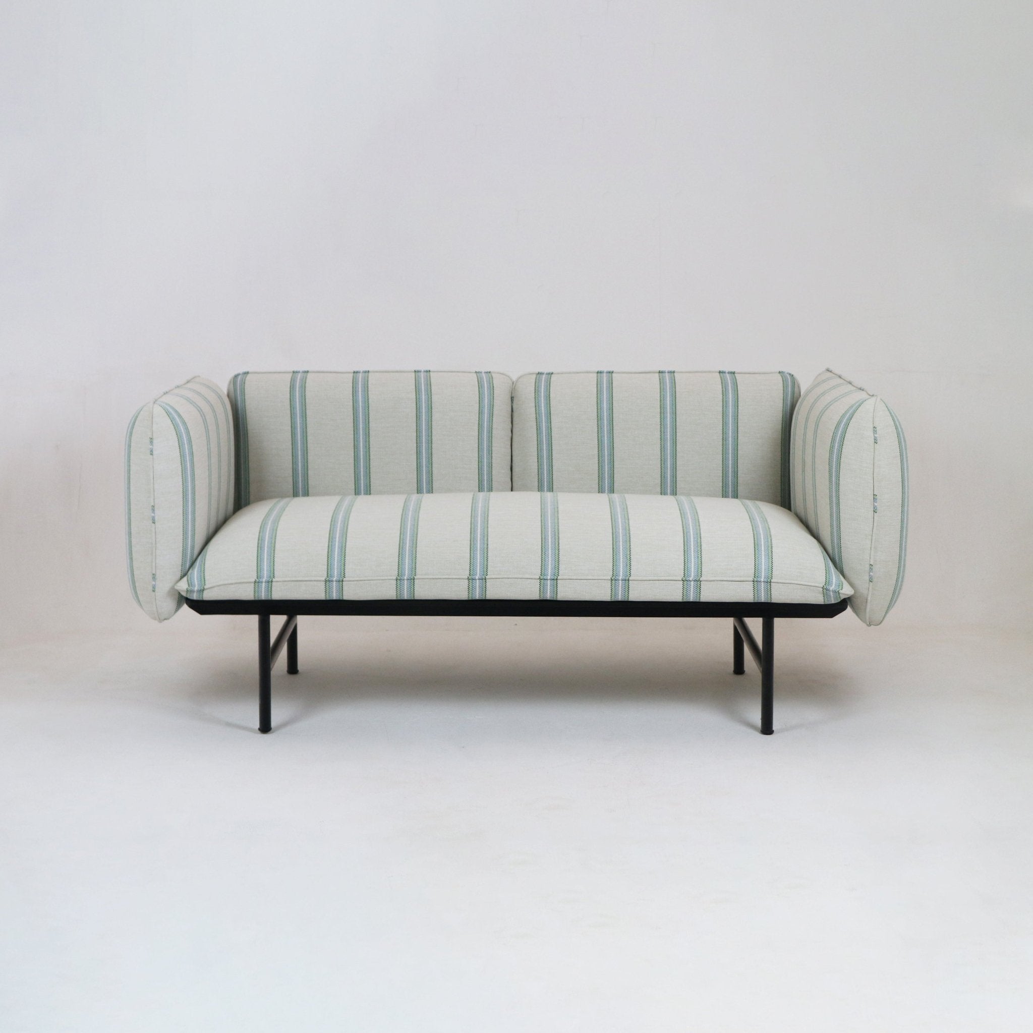 Coastal-inspired outdoor sectional sofa upholstered in light neutral striped fabric with pale green and blue vertical stripes, rounded padded arms and a low black metal frame and legs on a neutral studio background.