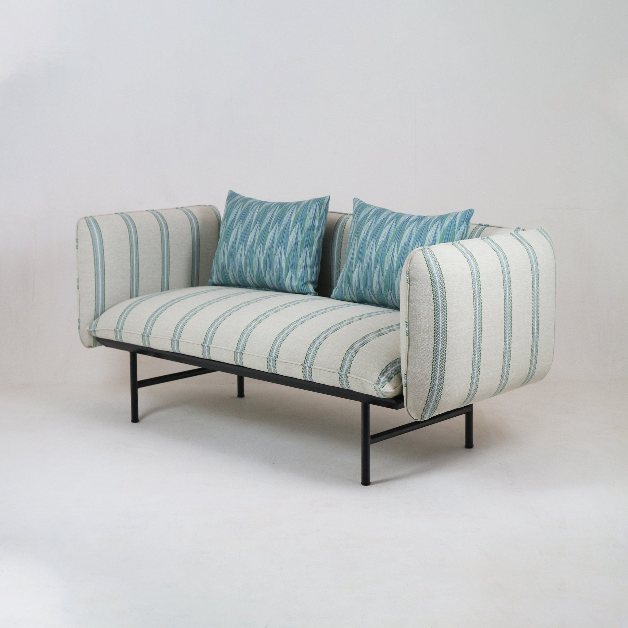 Coastal outdoor two-seat sectional sofa upholstered in cream fabric with pale blue-green stripes, styled with two blue-green patterned pillows, teak-accented details and a slim black metal frame with legs.