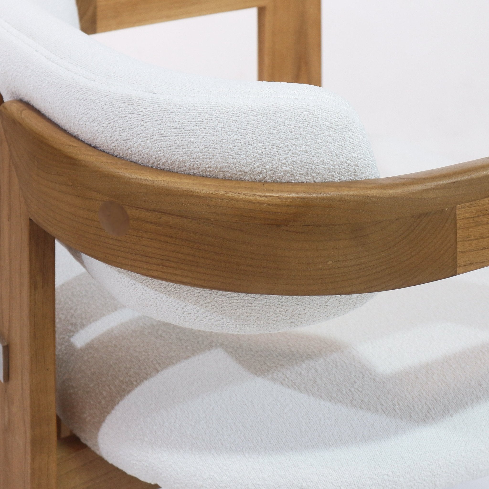 Close-up of a dining chair showing a curved teak frame and white boucle upholstery, highlighting the wood grain, round joinery plug, and the textured boucle seat and back cushion.