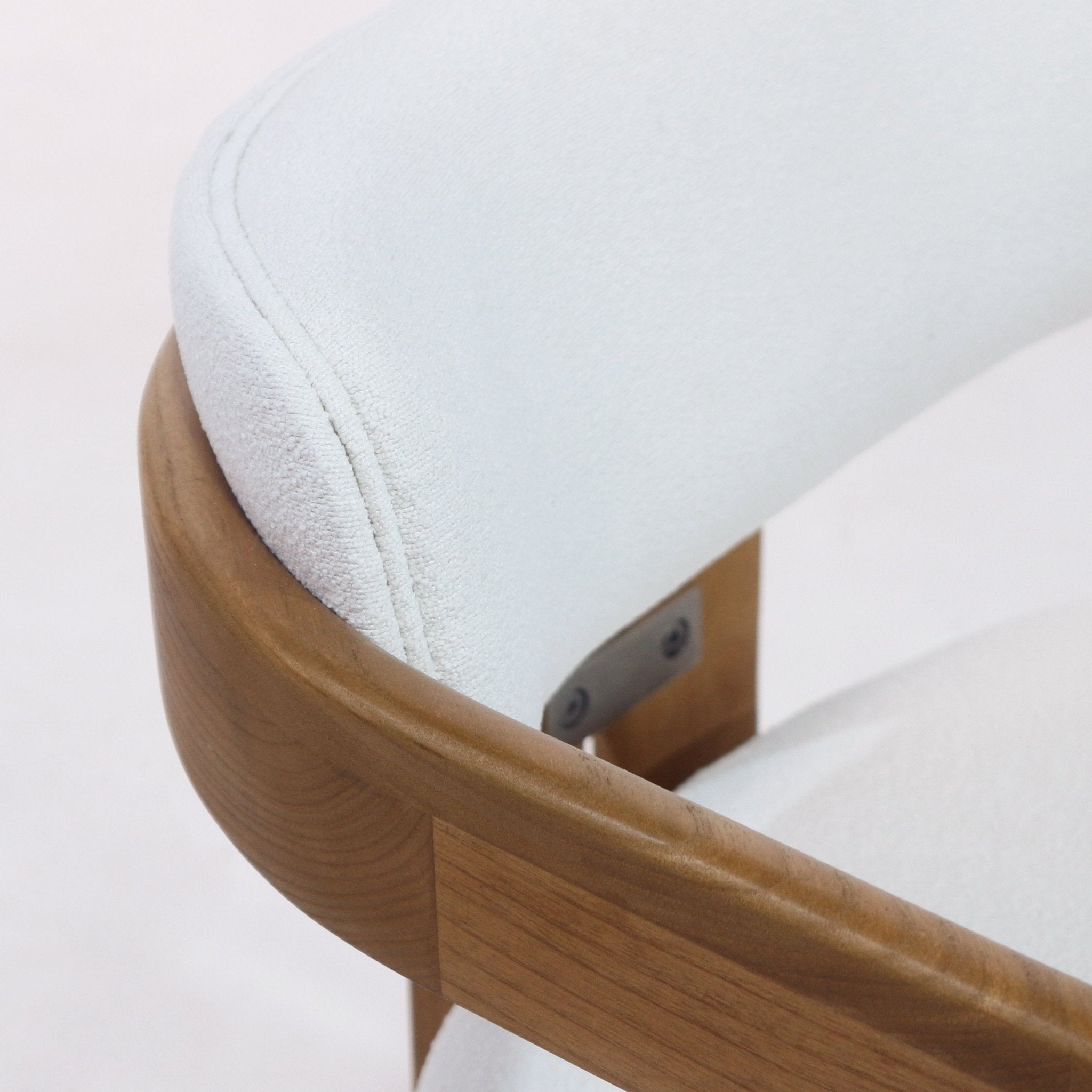 Close-up of a dining chair highlighting the curved teak wood arm and frame with white boucle upholstery on the back and seat, showing seam stitching and a metal bracket where the back connects to the frame.