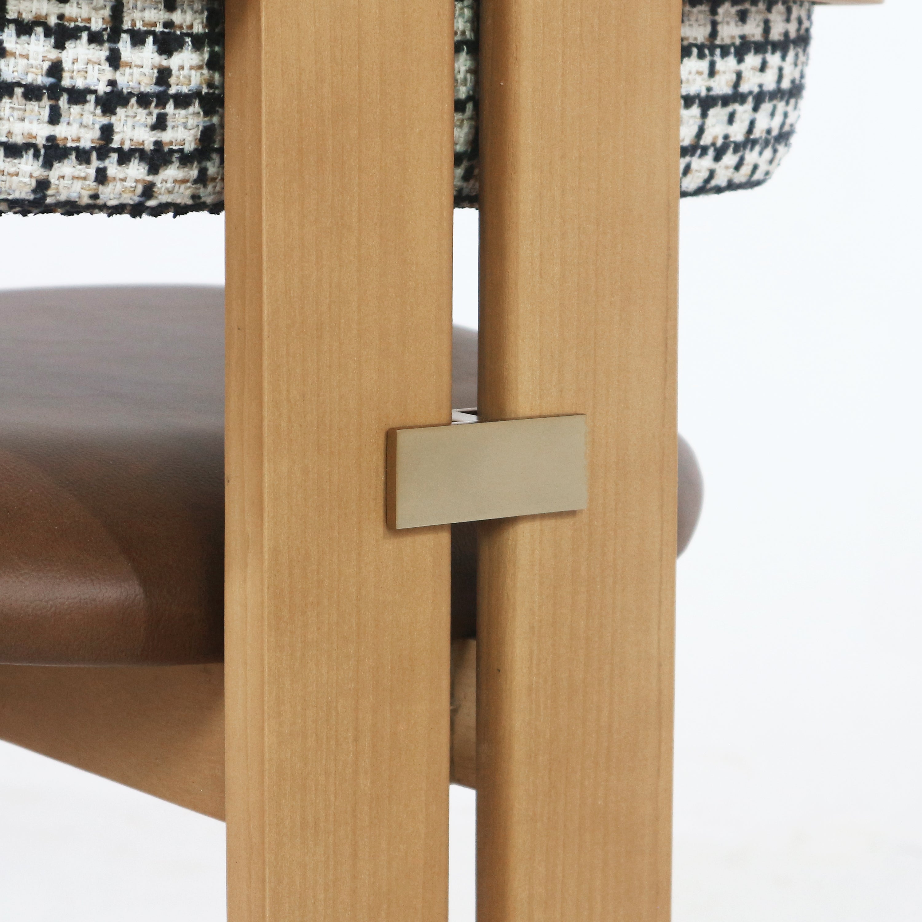 Close-up of a mid-century modern teak dining chair frame showing a rectangular brass connector between two light teak uprights, with a brown leather seat edge and black-and-white woven fabric back visible.
