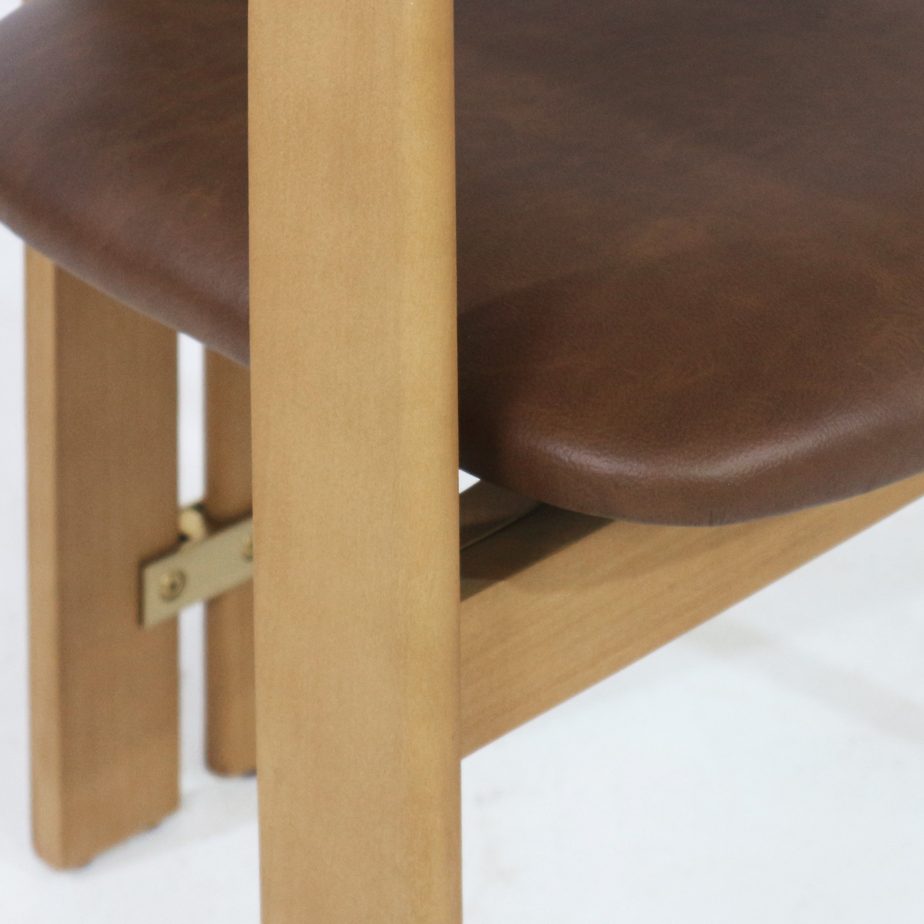 Close-up of a teak dining chair showing a light teak leg, rounded brown upholstered seat and a small brass support bracket beneath the seat.
