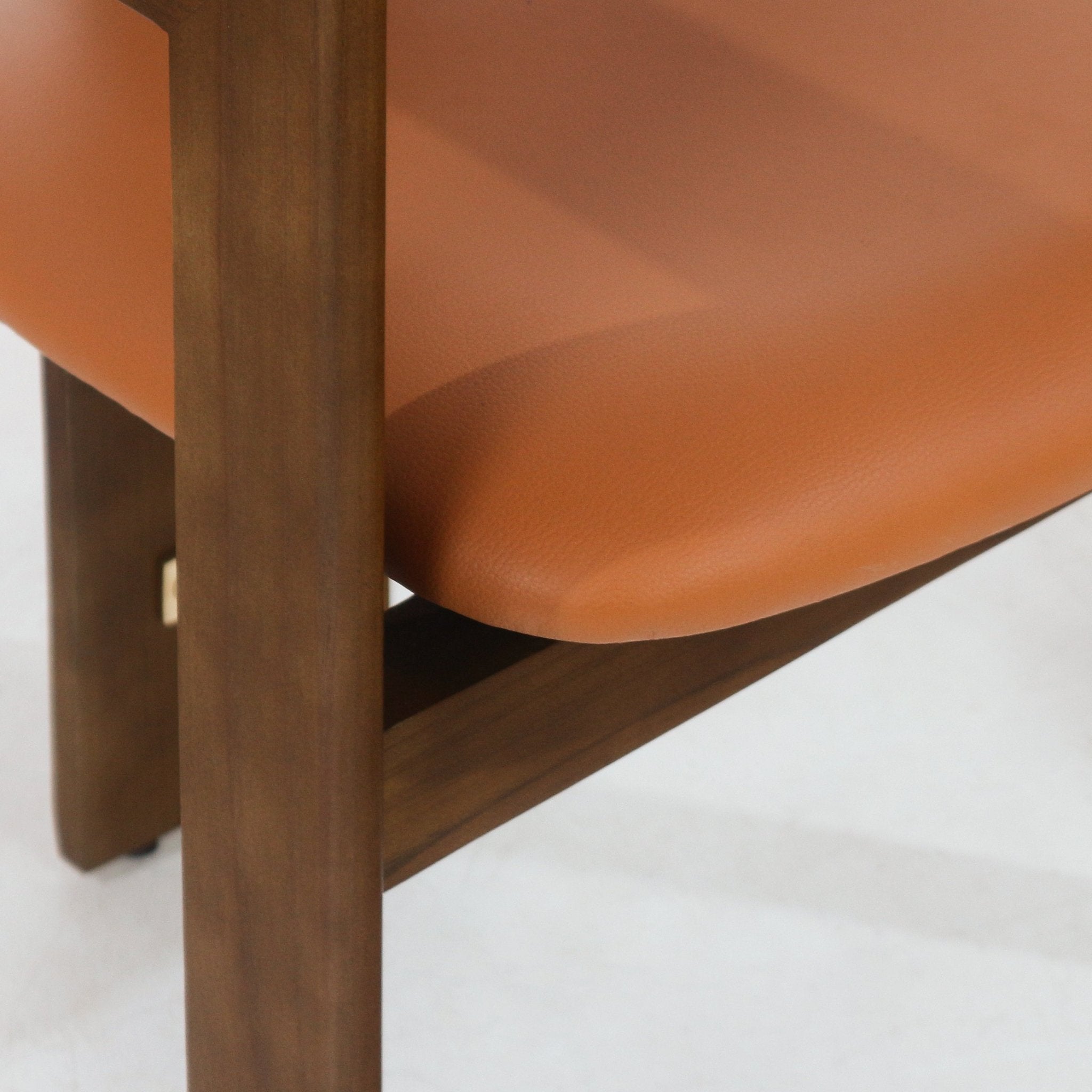 Close-up of a tan leather dining chair seat with a rounded padded edge resting on a walnut wood leg and diagonal support, highlighting the leather grain and warm walnut/teak mid-century modern finish.