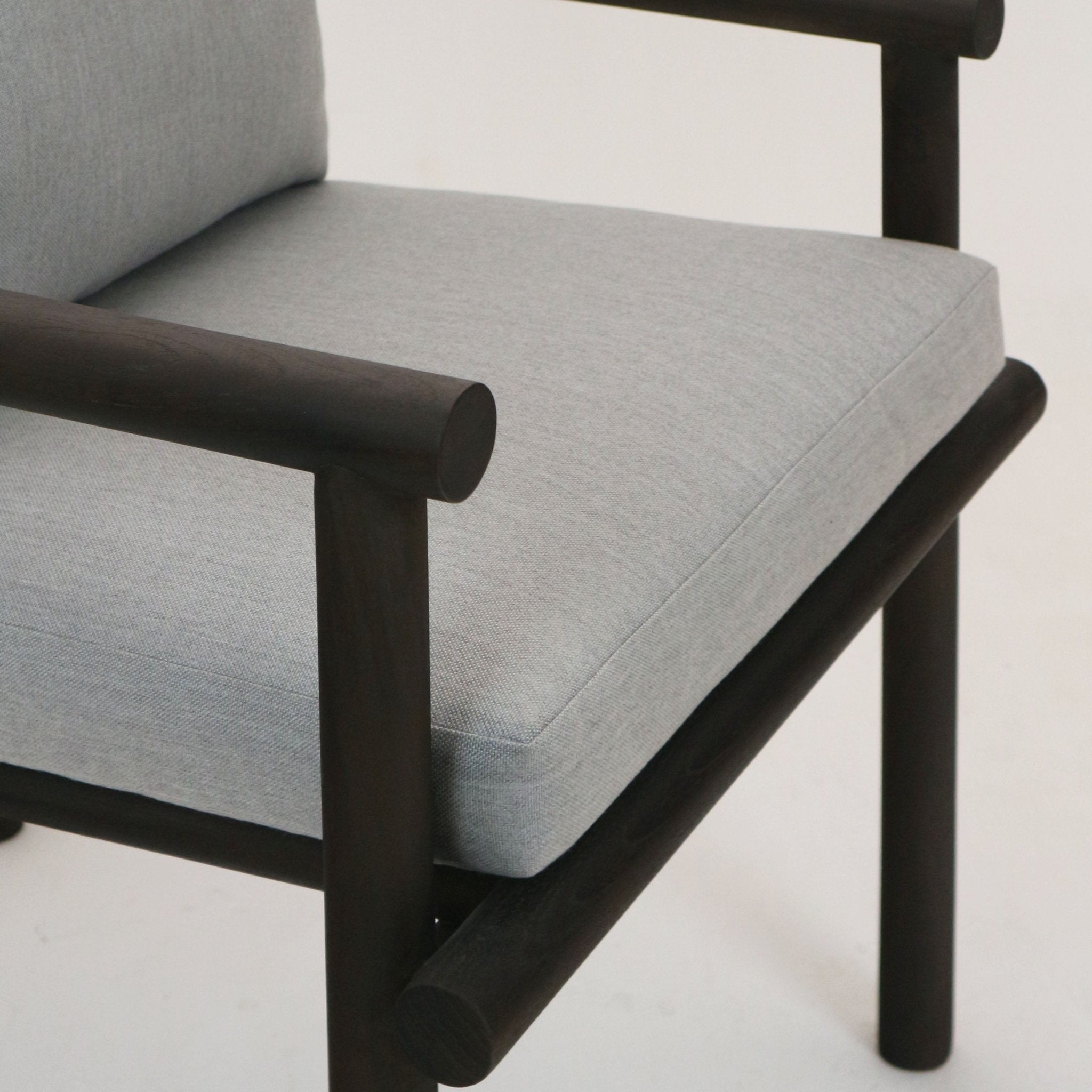 Close-up of a patio chair seat showing a light gray Sunbrella fabric cushion on a dark walnut-stained teak frame with rounded tubular arm and leg details.