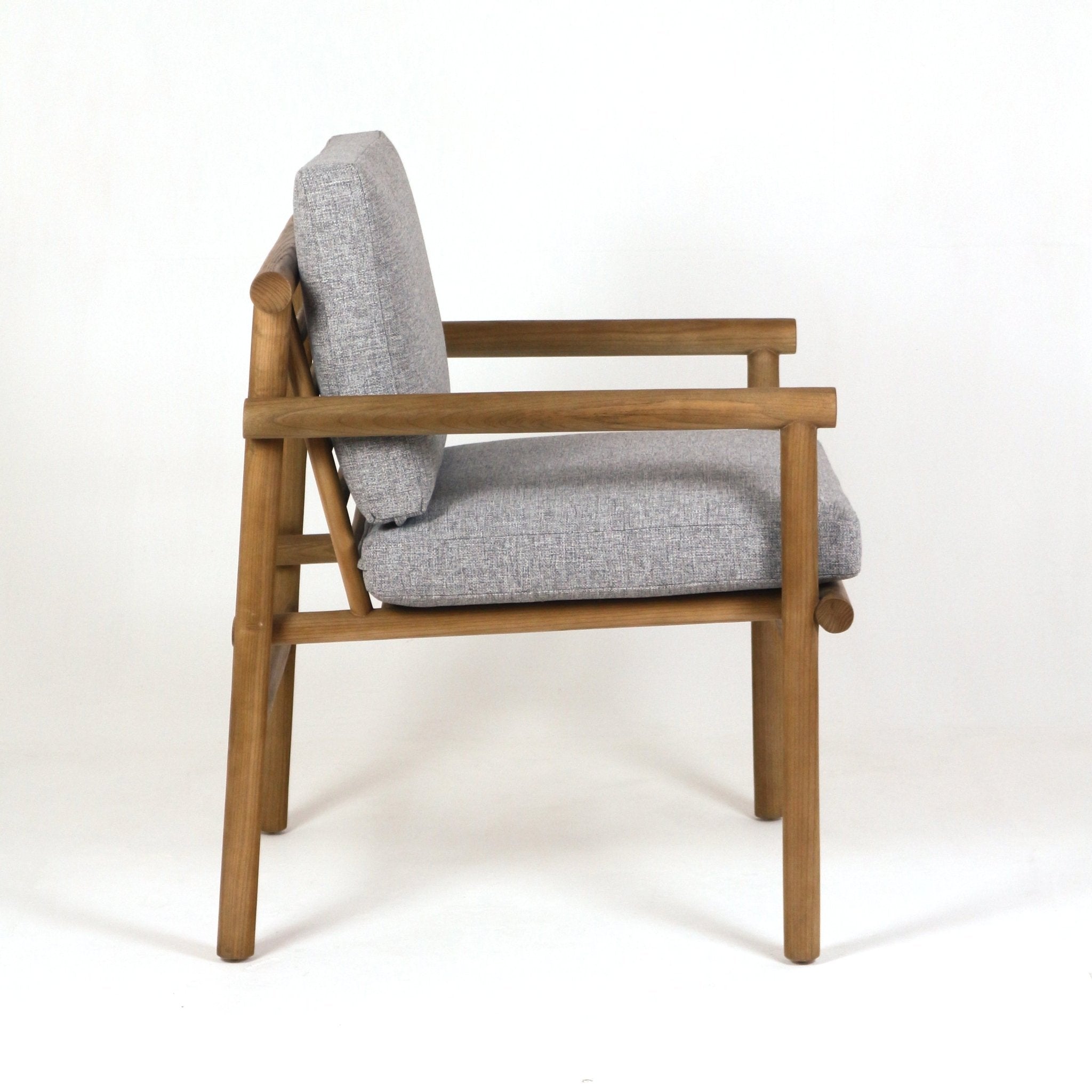 Side view of a natural teak patio chair with light gray performance fabric cushions, rounded wooden arms, ladder-style back supports and tapered legs on a white background.