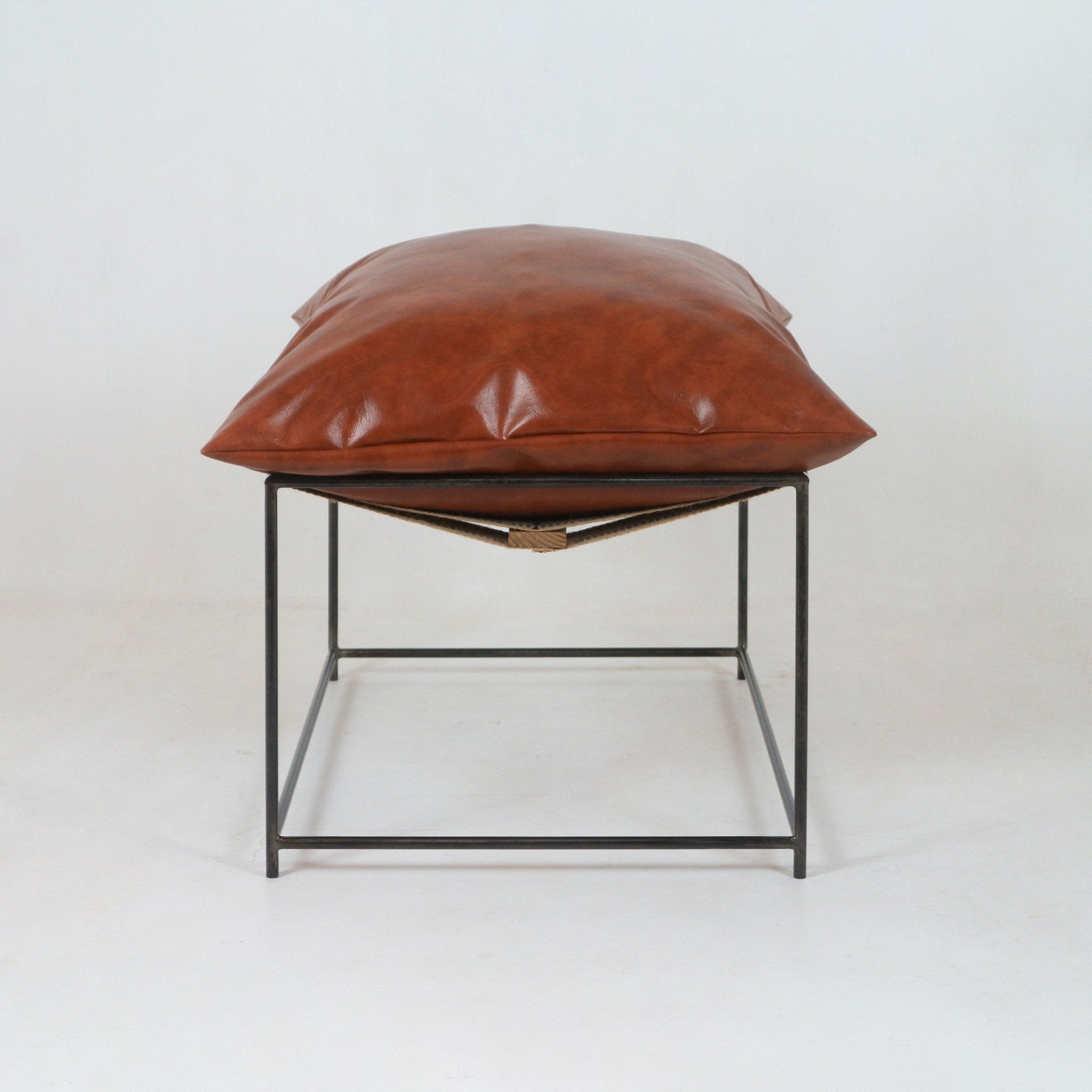 Modern plush red leather ottoman with a pillow-like cushion resting on a minimalist black metal frame and natural woven sling, photographed on a white studio background.