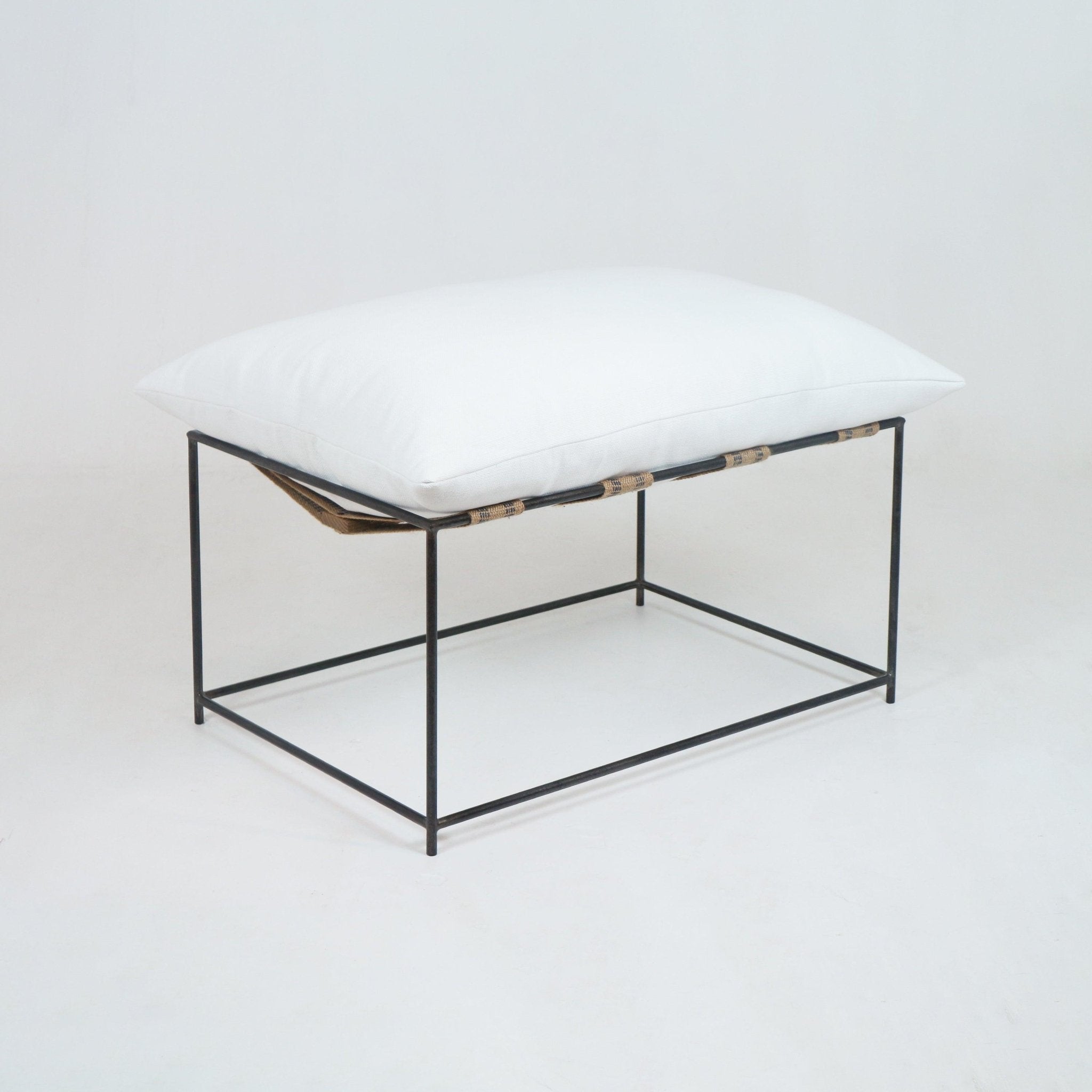 Modern ottoman with a plush white linen cushion resting on a minimal black steel frame with natural woven straps, photographed on a clean white background.