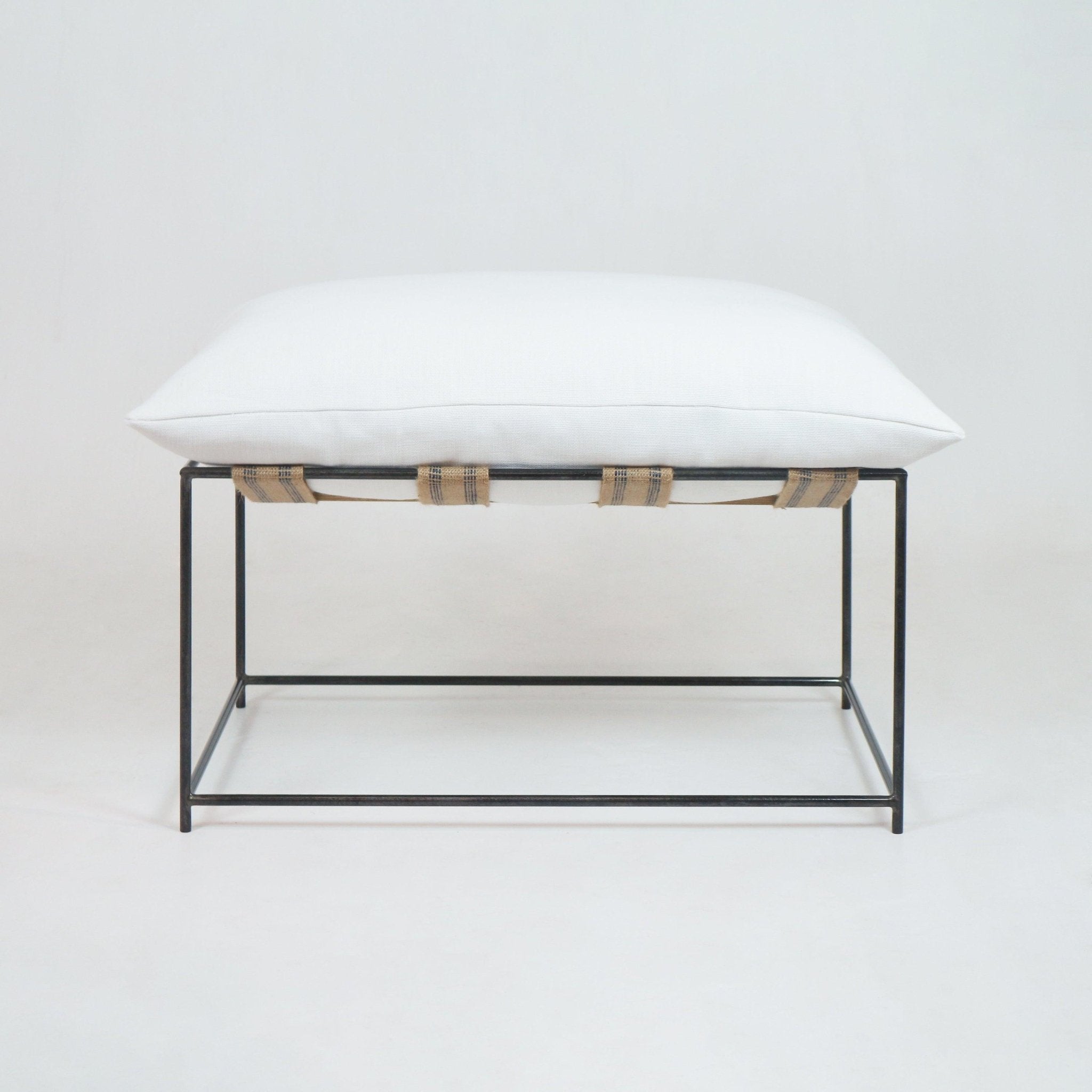Front view of a modern outdoor ottoman with a white linen cushion atop a slim dark metal frame, supported by natural woven straps, shown on a white studio background.