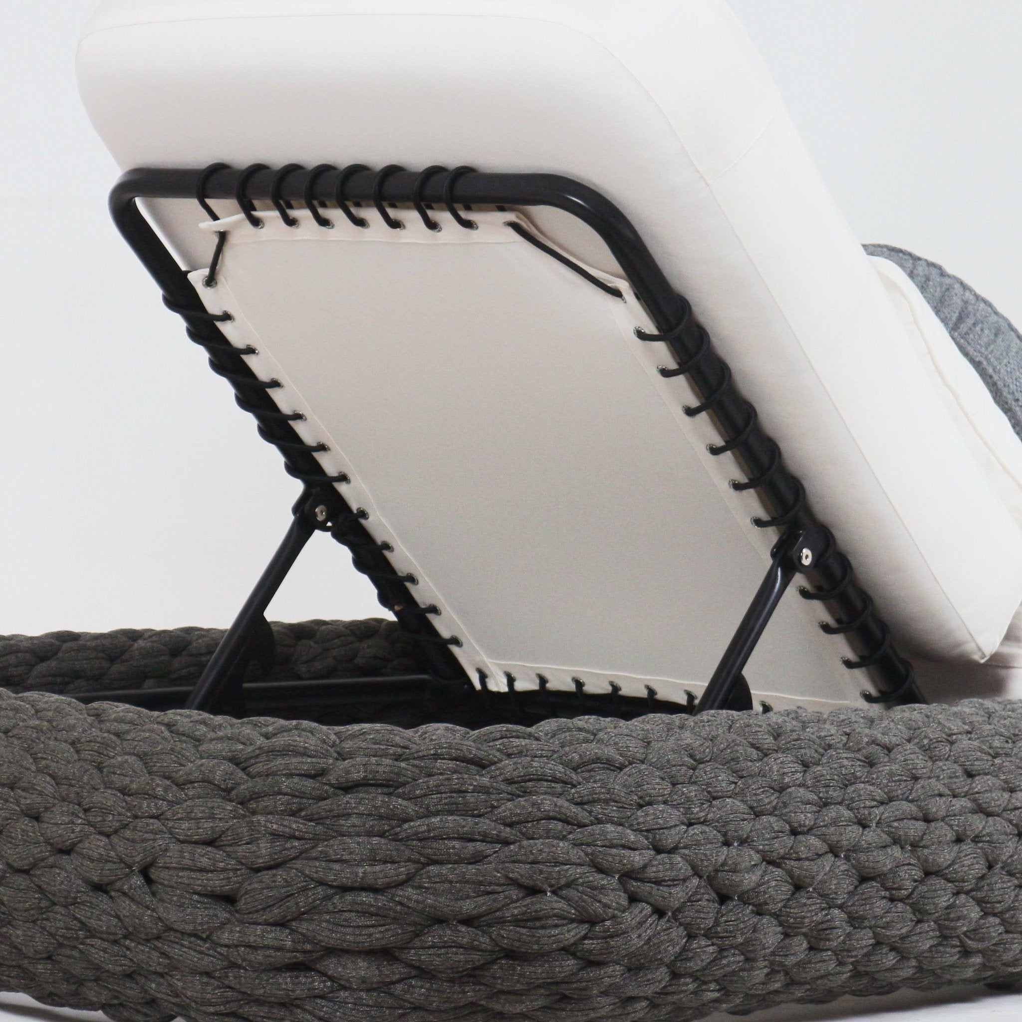 Close-up of a poolside chaise back showing a taupe fabric cushion attached to a black metal frame with laced rope ties and adjustable hinge, sitting on a dark braided rope-woven base.