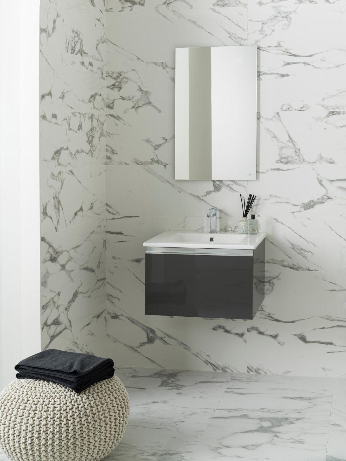 Modern floating 24.5-inch single-sink vanity with white stone top and integrated ceramic basin, dark gray solid-wood drawer, chrome faucet and rectangular mirror against marble-look tile walls and floor, knitted pouf with towels in foreground.
