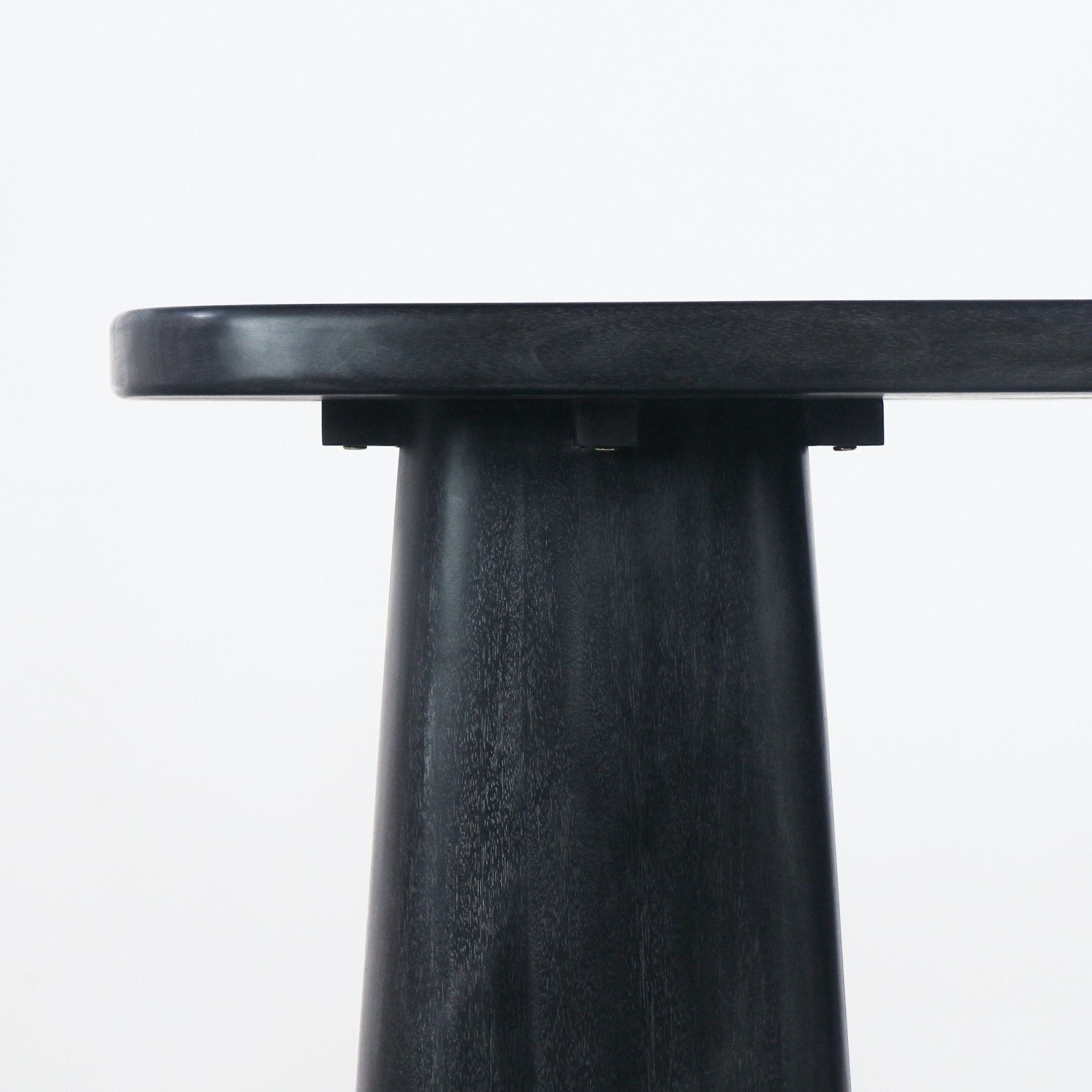 Close-up of a smoked black solid wood console showing the rounded tabletop edge and tapered pedestal leg with visible wood grain, a modern minimalist detail against a white background.