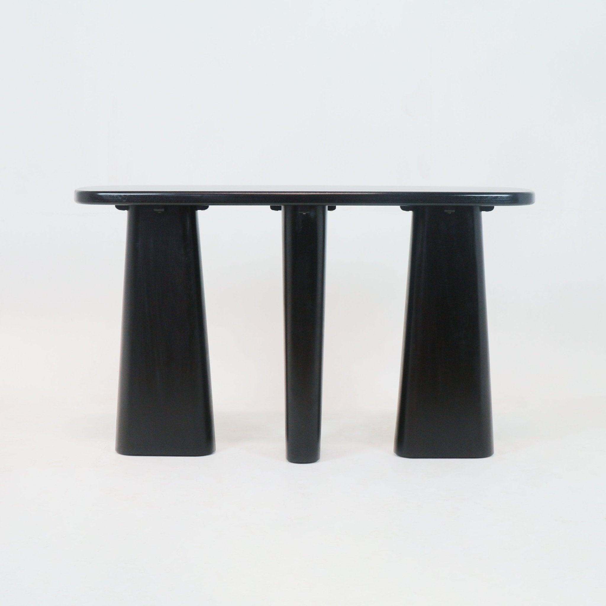 Smoked-black solid wood console table with a rounded rectangular top and three pedestal legs—two flared trapezoid outer legs and a central round leg—in a minimalist modern style on a white background.