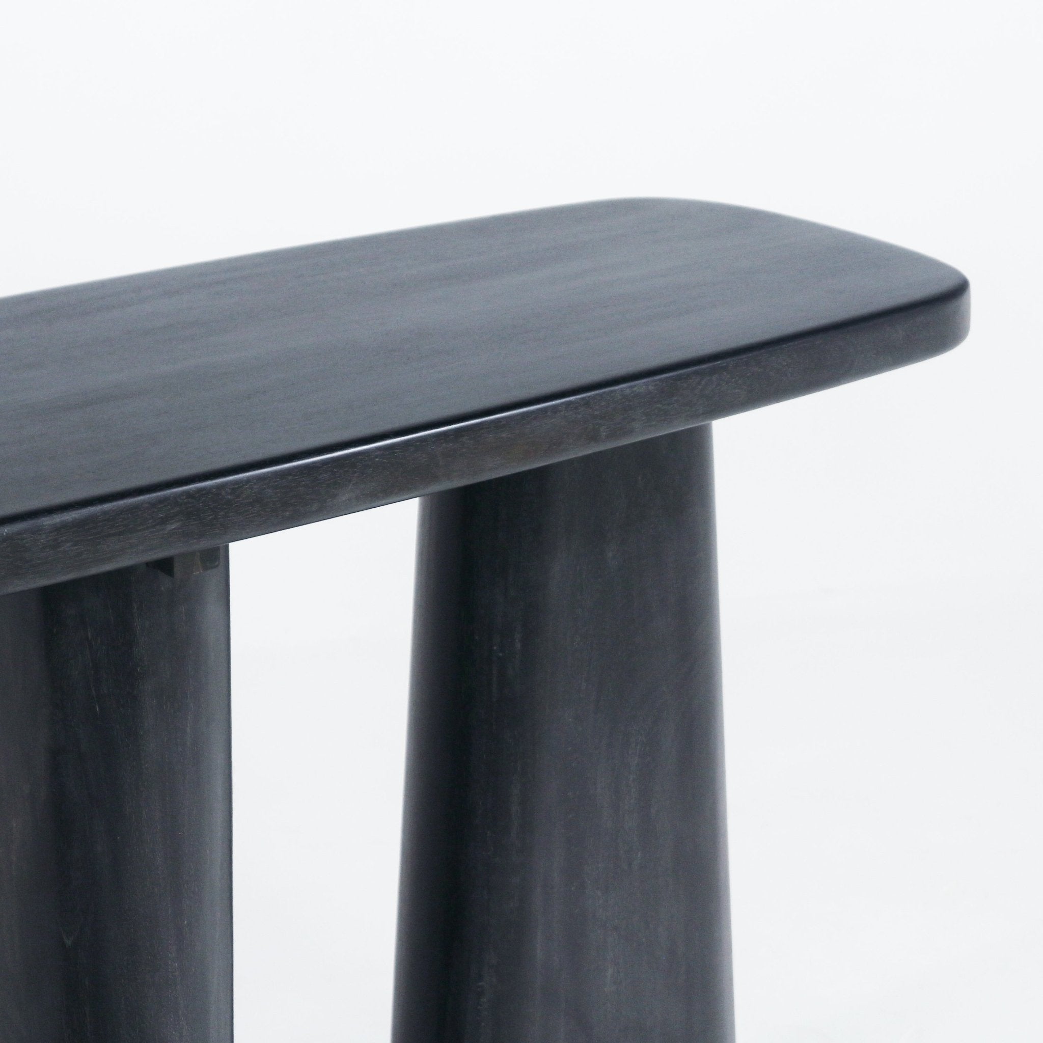 Close-up of a smoked-black solid wood console showing the rounded rectangular tabletop edge and two tapered cylindrical legs with a smooth, slightly weathered black finish.