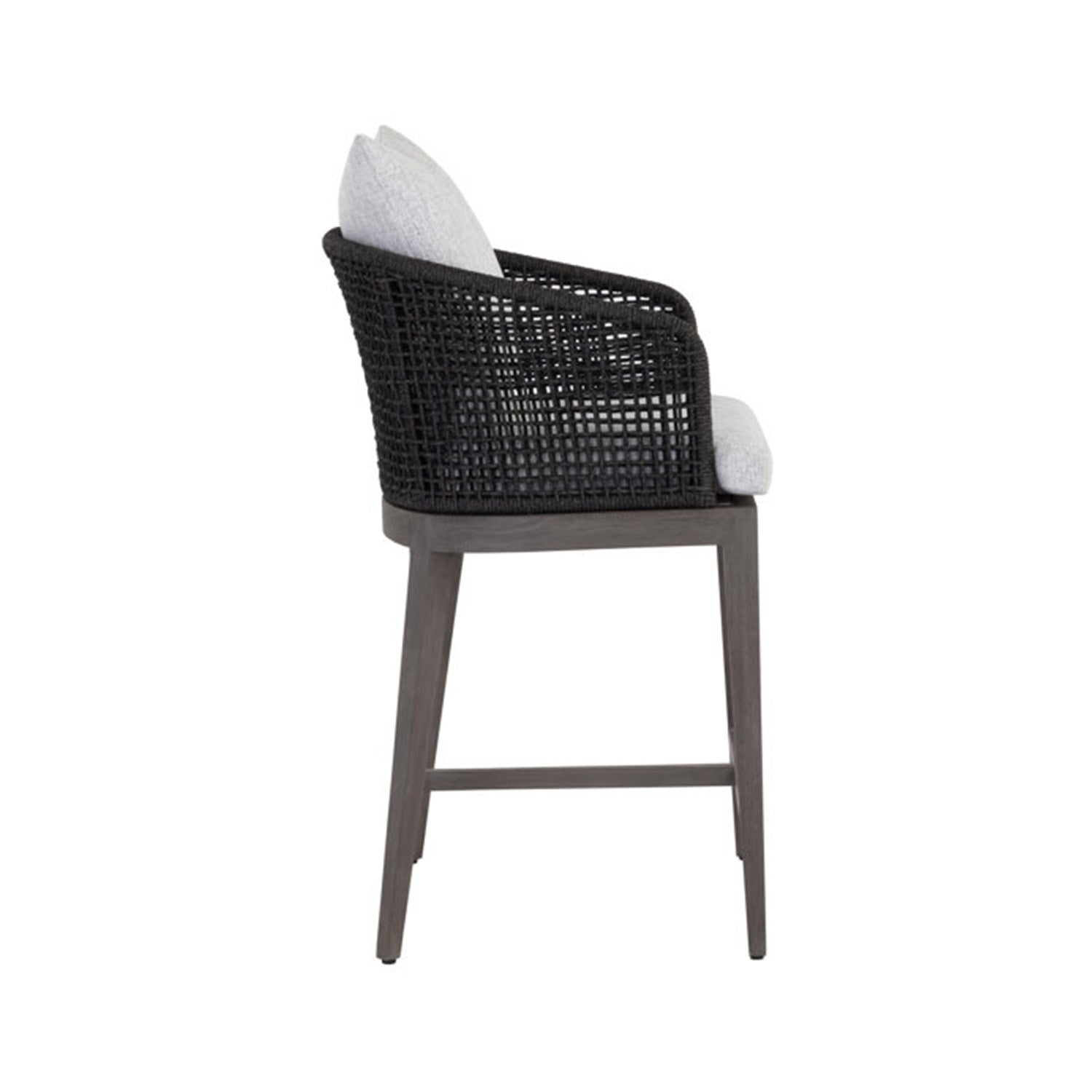 Side view of a teak outdoor bar stool featuring a dark woven rope back, light gray upholstered seat and cushion, tapered teak wood legs and a built-in footrest.