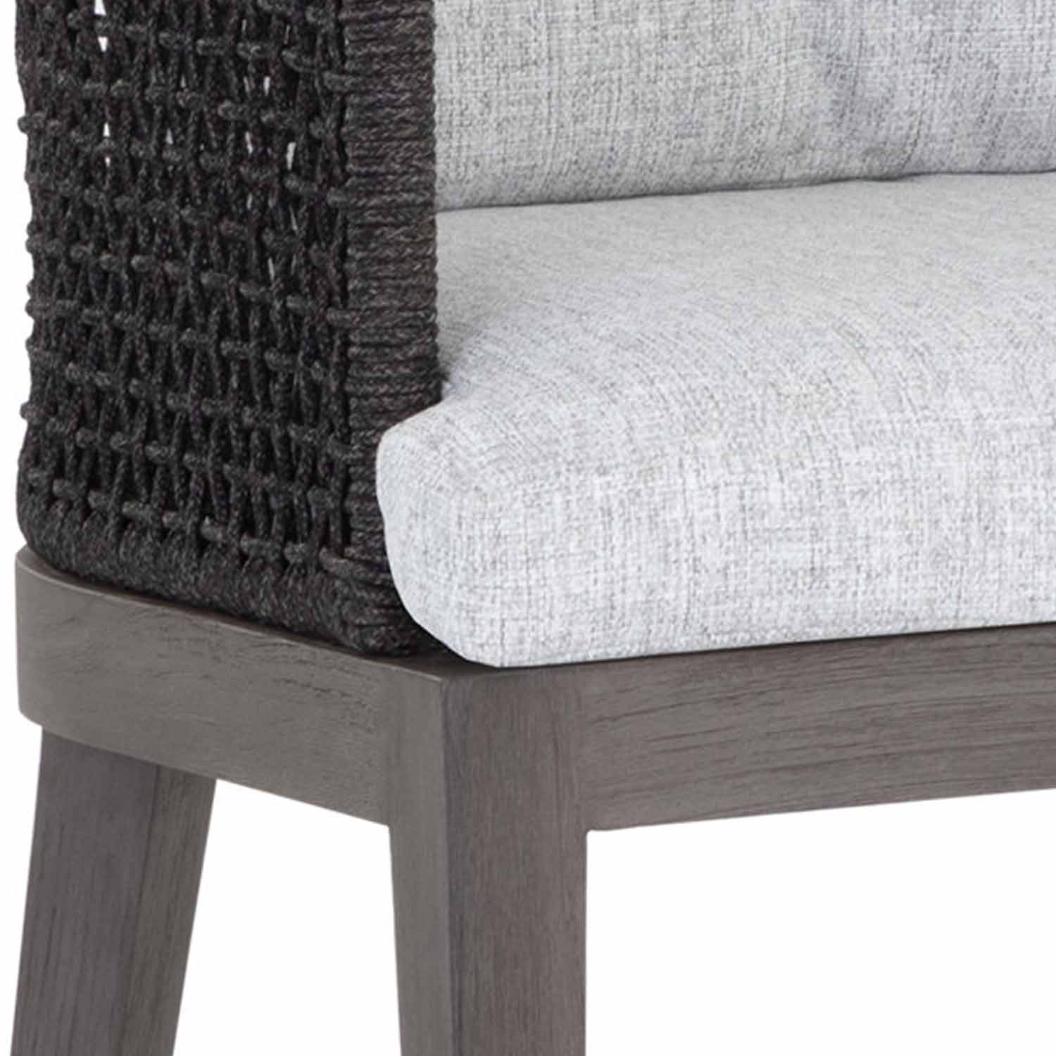 Close-up of an outdoor teak bar stool corner showing a gray-stained teak wood frame, woven dark rope back detail and a light gray upholstered fabric seat cushion.
