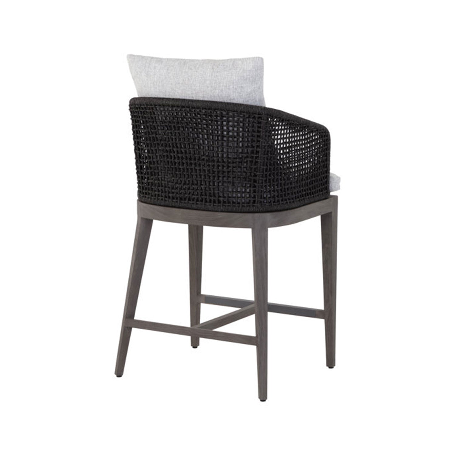 Rear three-quarter view of a teak outdoor bar stool with a black woven back, gray upholstered seat cushion and pillow, and tapered natural wood legs with a crossbar footrest.