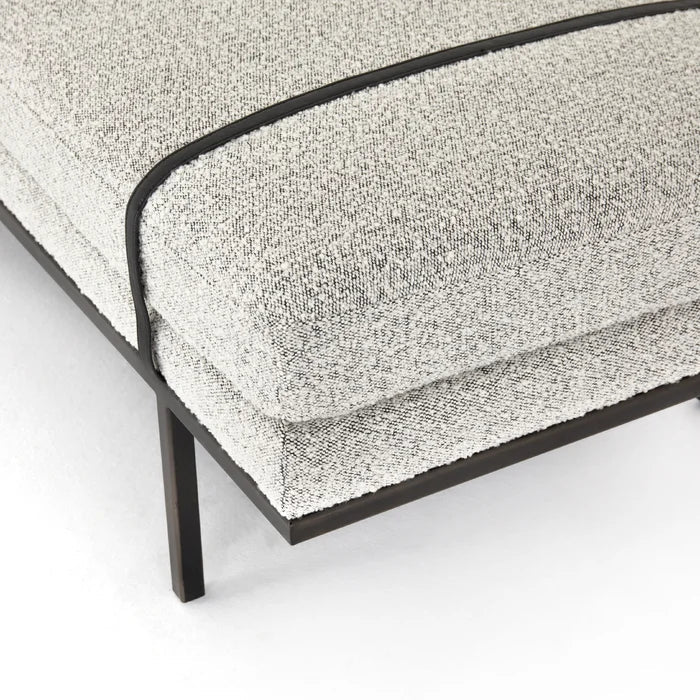 Close-up of a natural fabric boucle bench corner showing textured off-white boucle upholstery, contrasting dark piping and a slender dark frame leg, styled for a modern coastal bedroom.