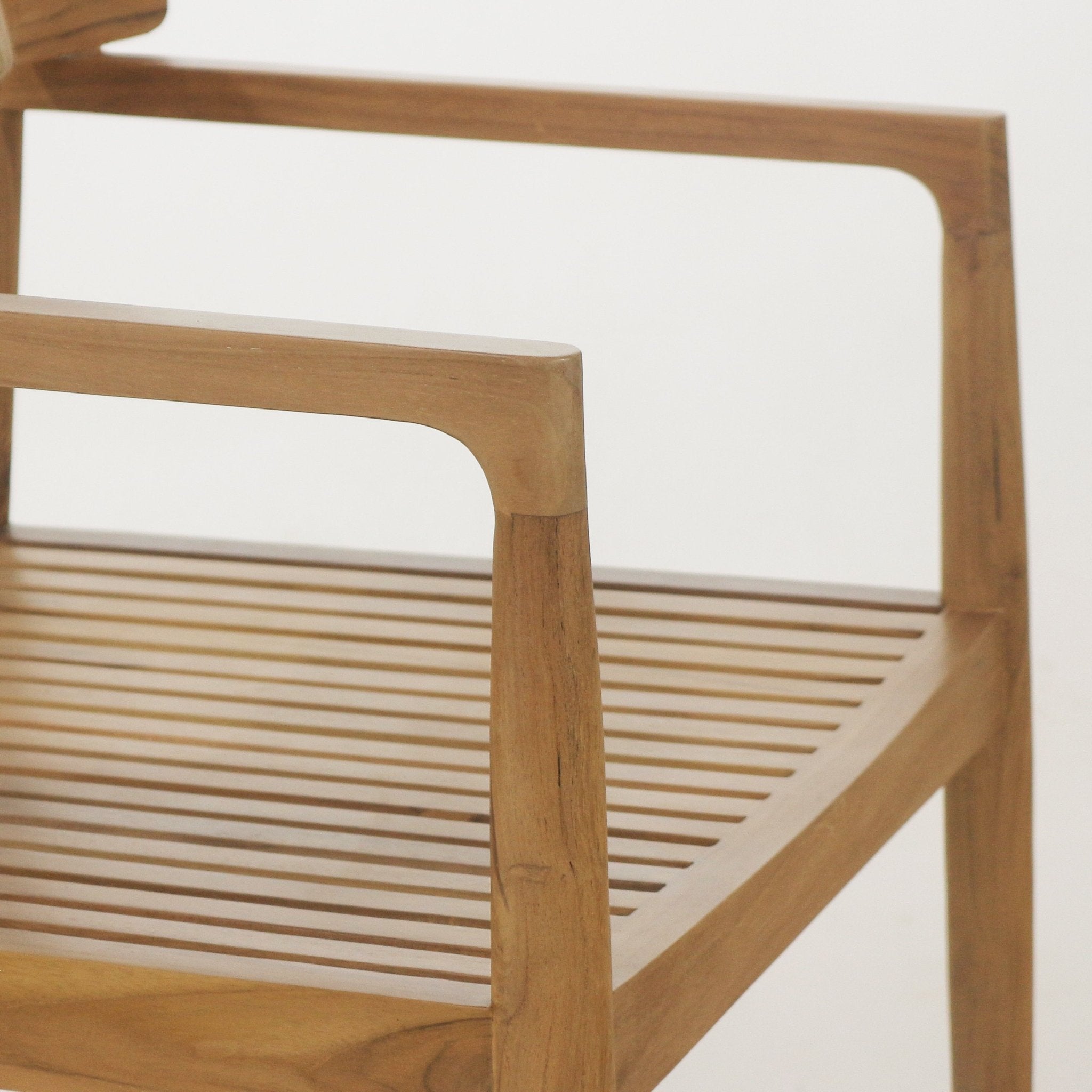 Close-up of an outdoor teak dining chair arm and slatted seat showing natural brown teak wood grain, smooth rounded armrest and tapered leg in a minimalist coastal/mid-century modern style.