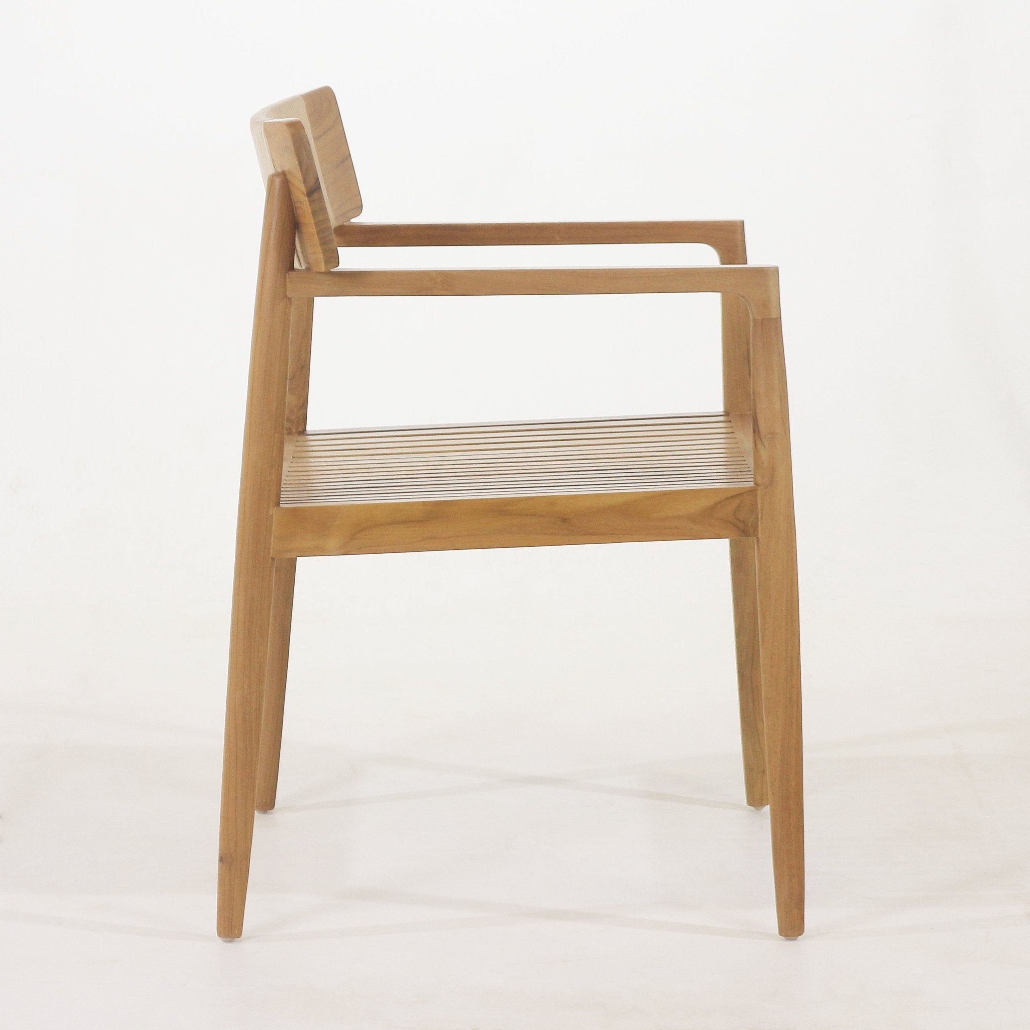 Side view of a natural teak outdoor dining chair with slatted wooden seat, tapered legs, square backrest and slim armrests in a coastal mid-century modern style.