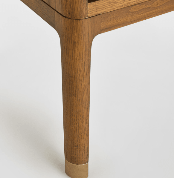 Close-up of a tapered solid-wood nightstand leg in warm brown oak grain with a brass toe cap, showing smooth rounded joinery and the underside of the bedside shelf.