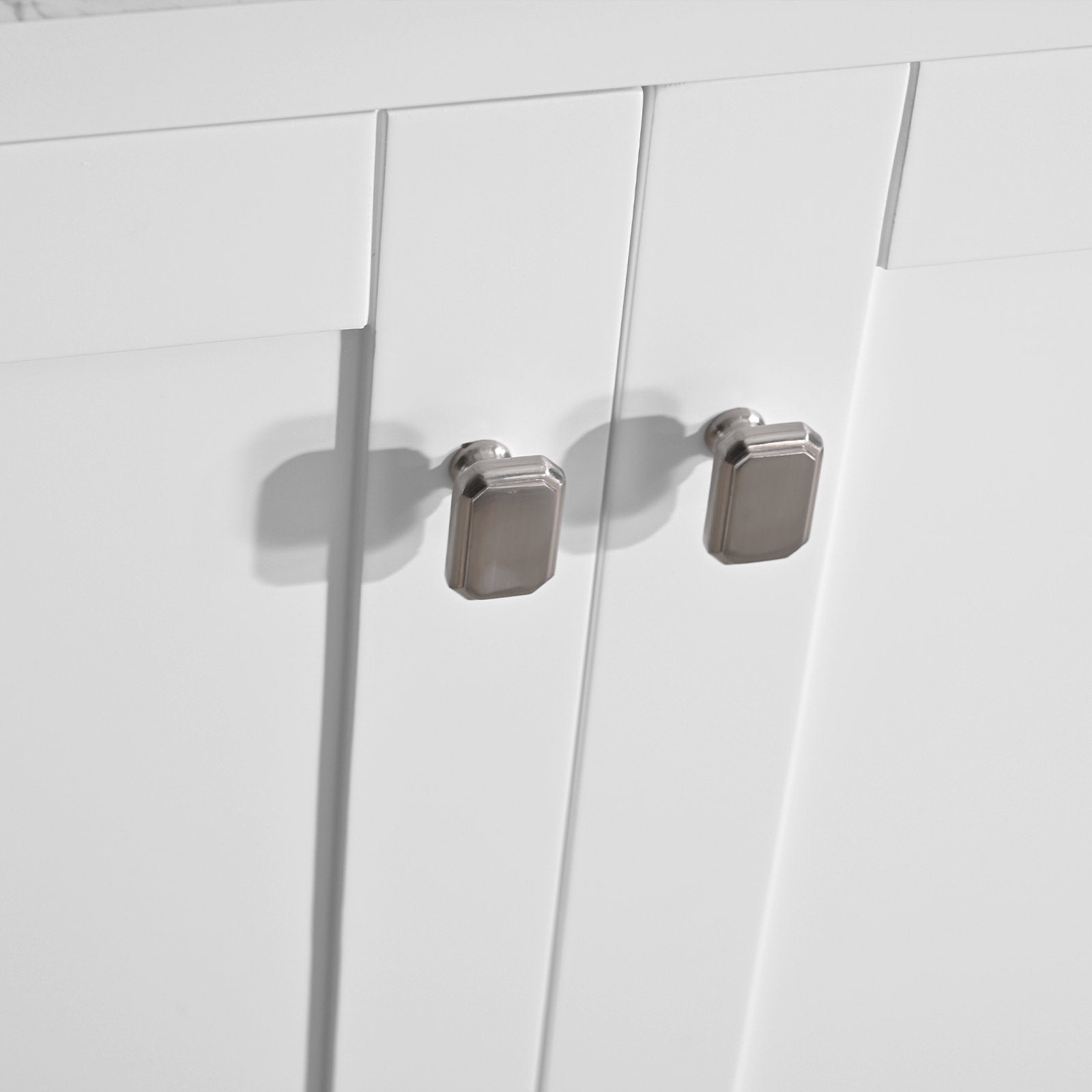 Close-up of white solid-wood vanity doors with a slim stone top edge, featuring two brushed-metal rectangular knobs and recessed panel detail casting soft shadows.