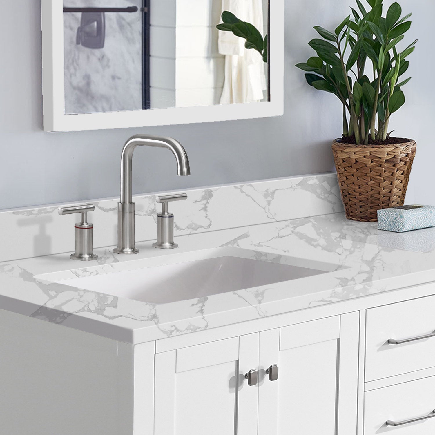 Freestanding white solid-wood bathroom vanity with a marble-look stone countertop, integrated undermount sink and brushed nickel faucet, styled with a wicker potted plant and rectangular mirror above.