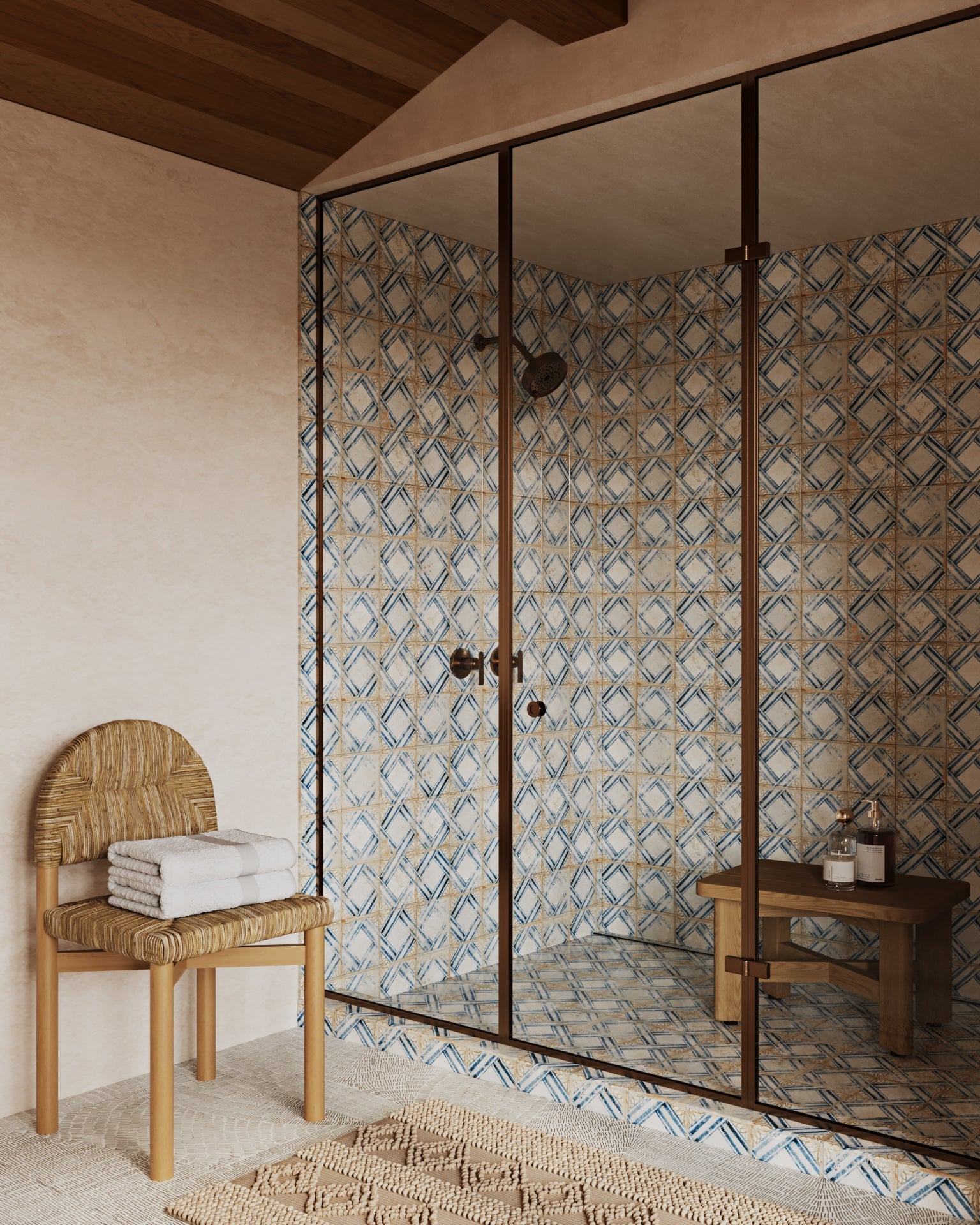 Teak shower stool and small teak bench sit inside a glass-enclosed shower with blue-and-beige patterned tile; a woven natural wood chair with folded white towels stands outside on a textured rug.