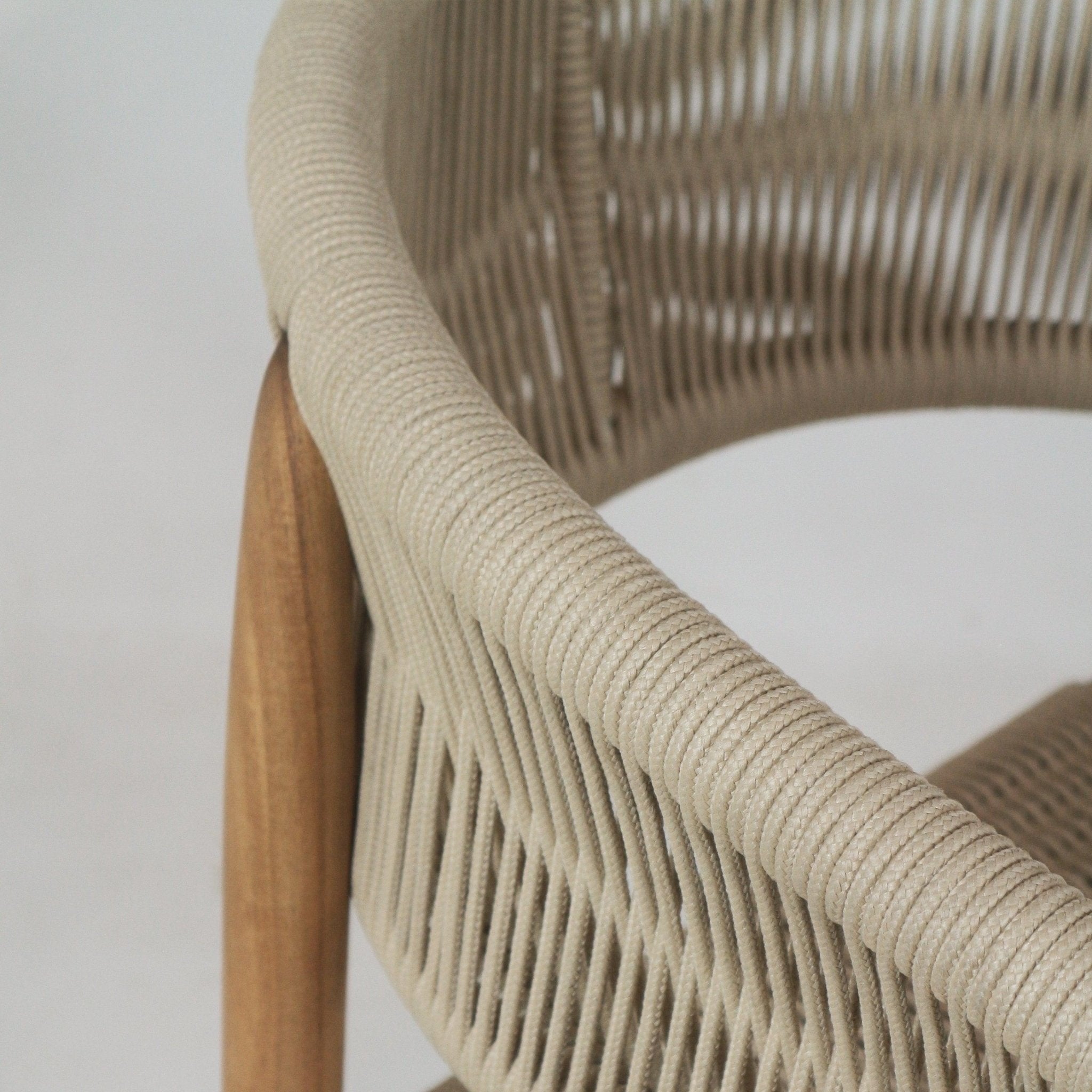 Close-up of a teak dining chair back showing a warm teak wood leg and beige polypropylene rope woven around the curved backrest and arm, highlighting the tight braided texture and coastal-modern design.